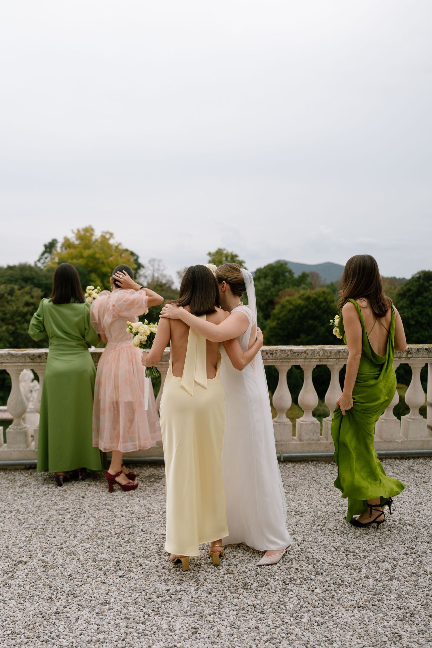 Wedding party in green and pink dresses pose with their backs turned on a stone balcony overlooking mountains.