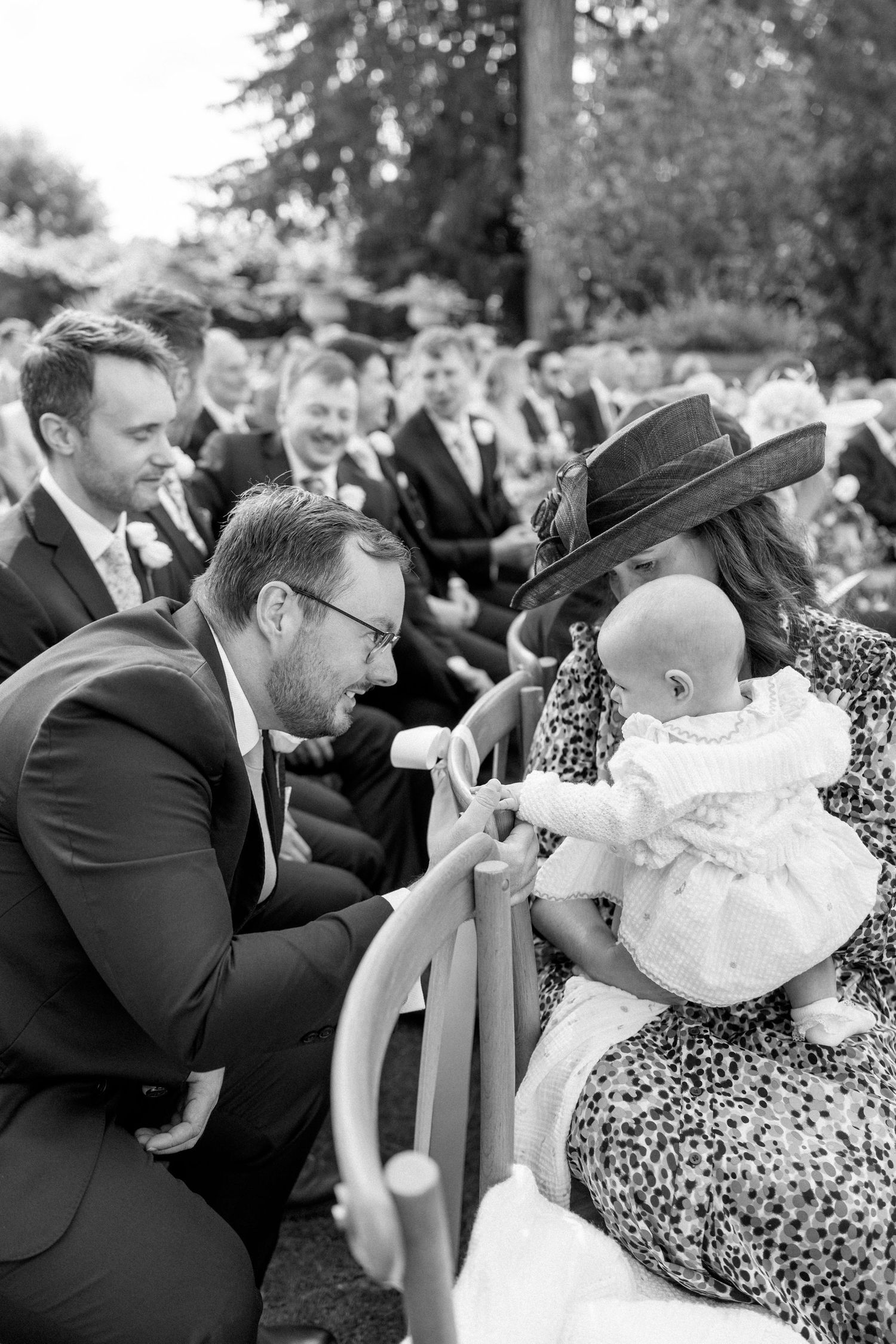 Wedding guest in suit interacts with baby wearing christening gown during outdoor ceremony.