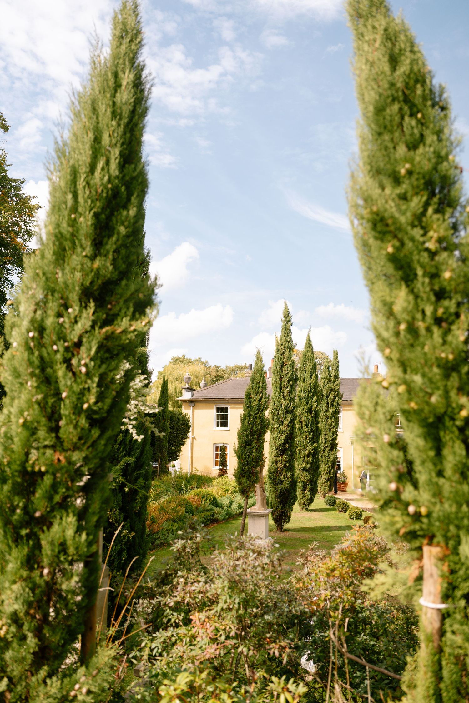 Tall cypress trees line a garden path against a blue sky and stone building.