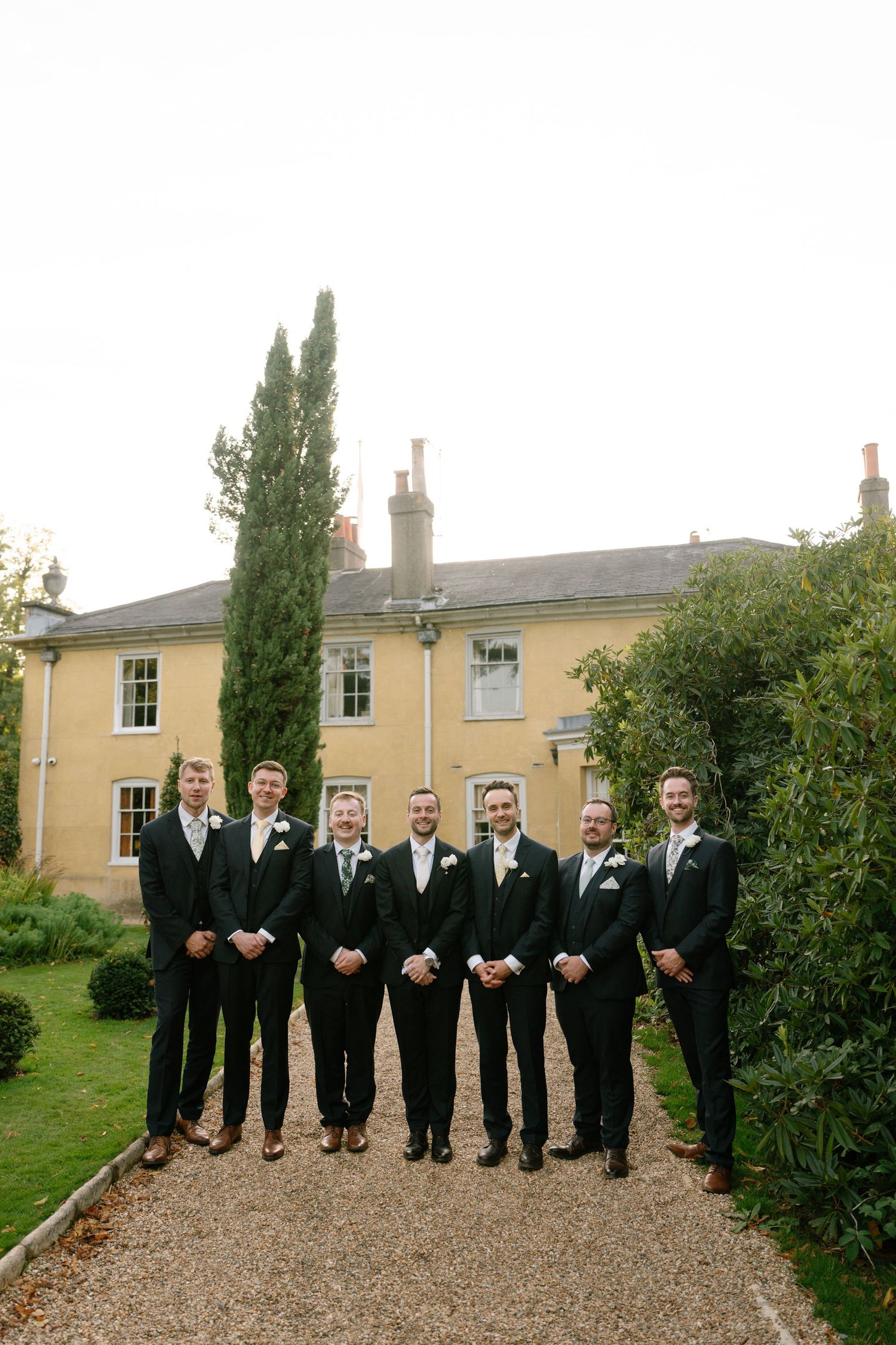 Formal group photo of groomsmen in black suits lined up outside a yellow manor house.