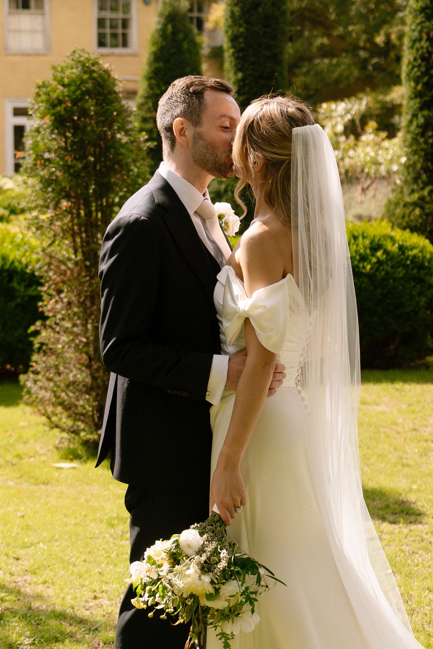 Romantic wedding moment captured in a sunny garden with yellow manor house in background.