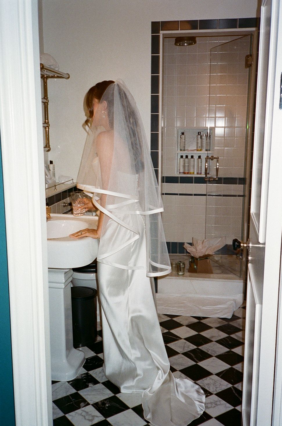 Bride in white wedding gown and veil standing at bathroom sink with black and white checkered floor.