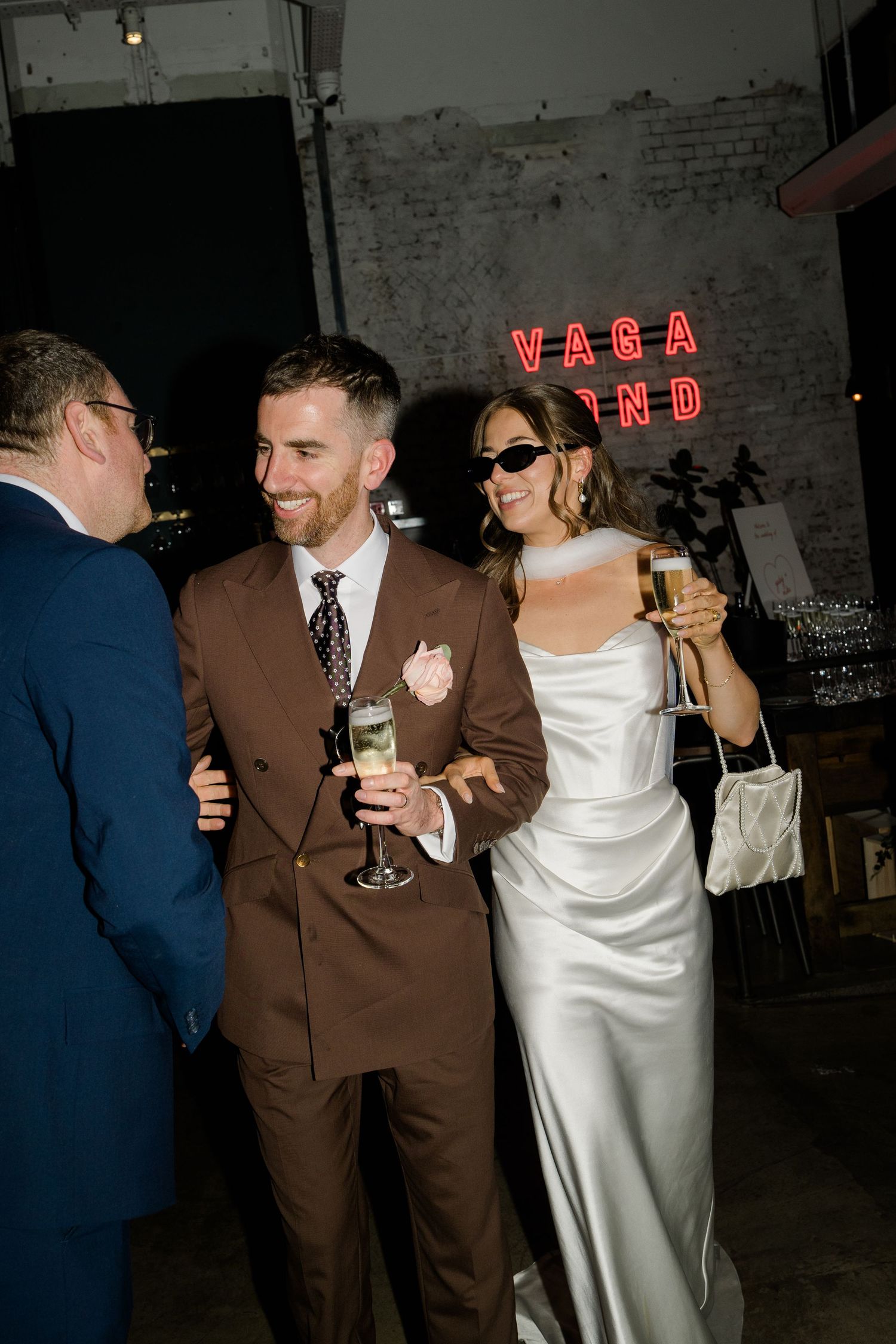 Bride in white satin dress and groom in brown suit celebrate with champagne near Vagabond neon sign.