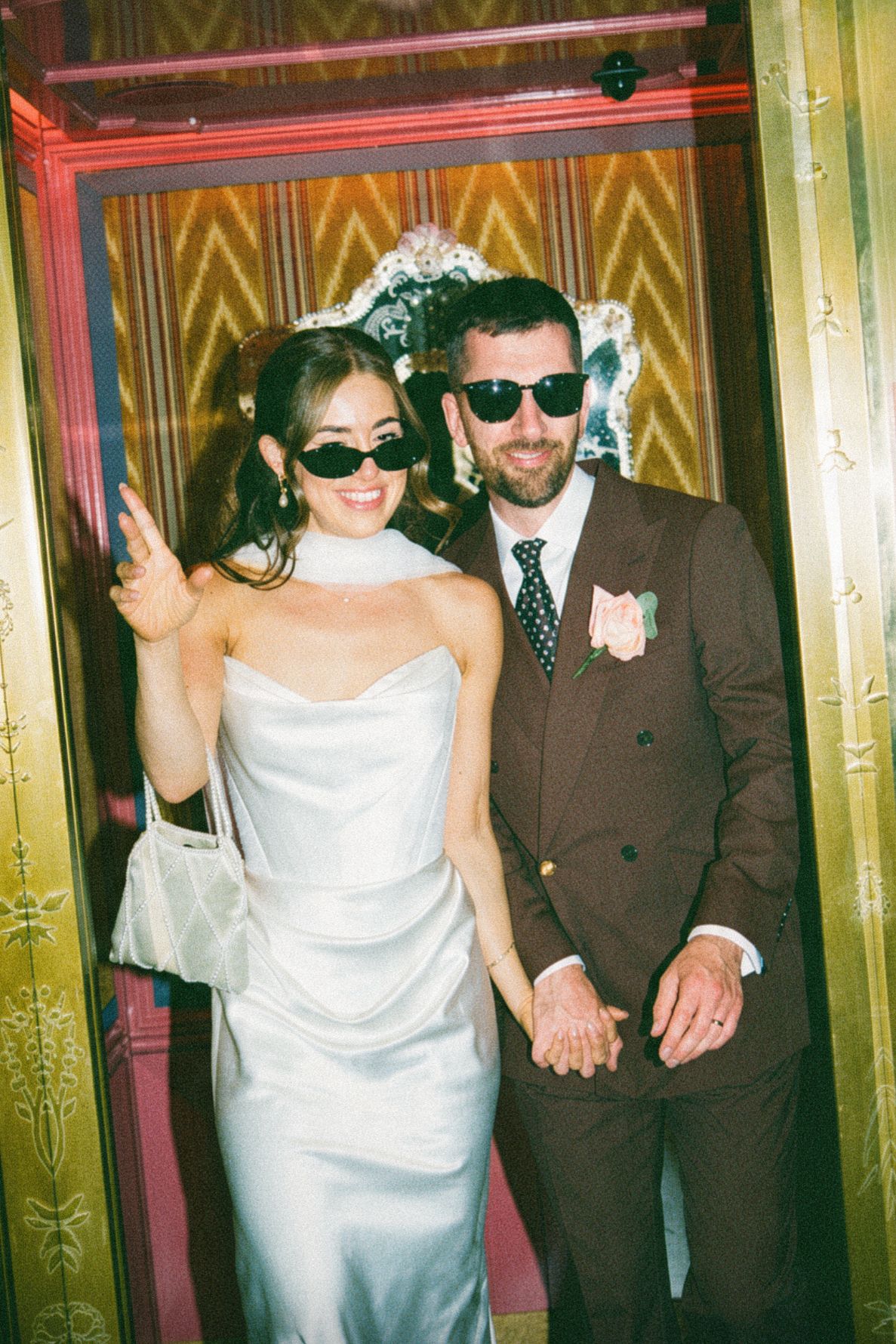 Stylish bride and groom wearing sunglasses holding hands in a gold art deco elevator at their wedding.