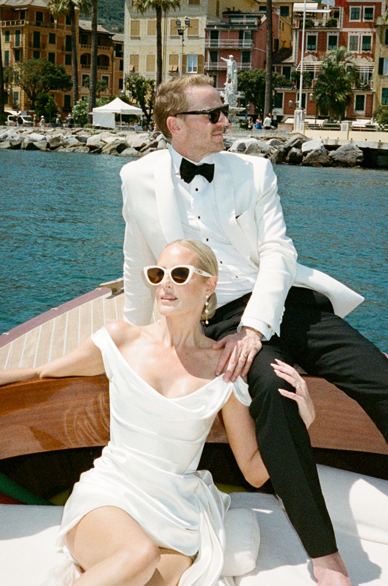 Bride in white gown and groom in white tuxedo pose elegantly on a wooden boat in Portofino harbor.
