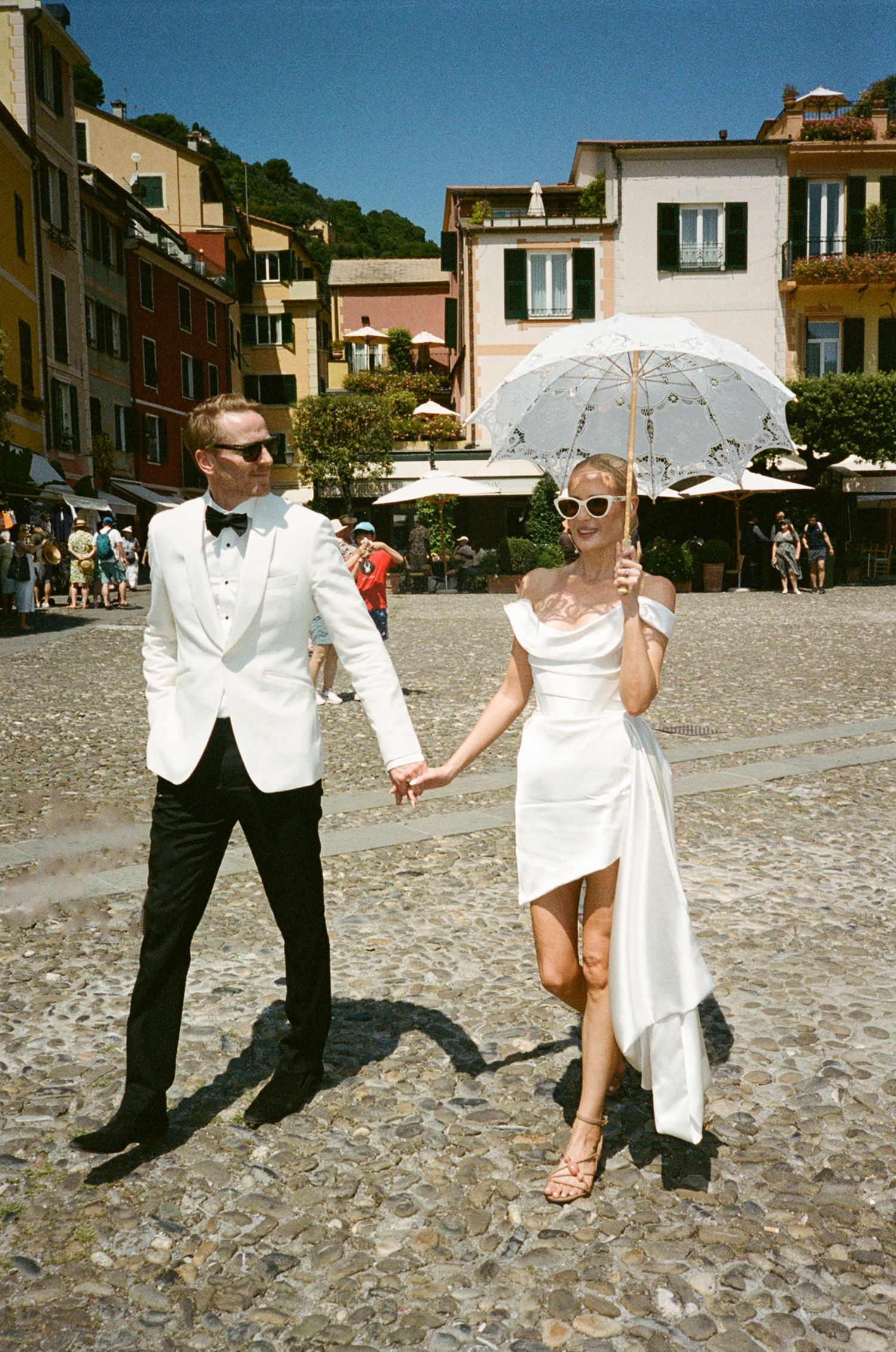 Bride and groom holding hands in white wedding attire on cobblestone plaza in Portofino, Italy, on a sunny day.