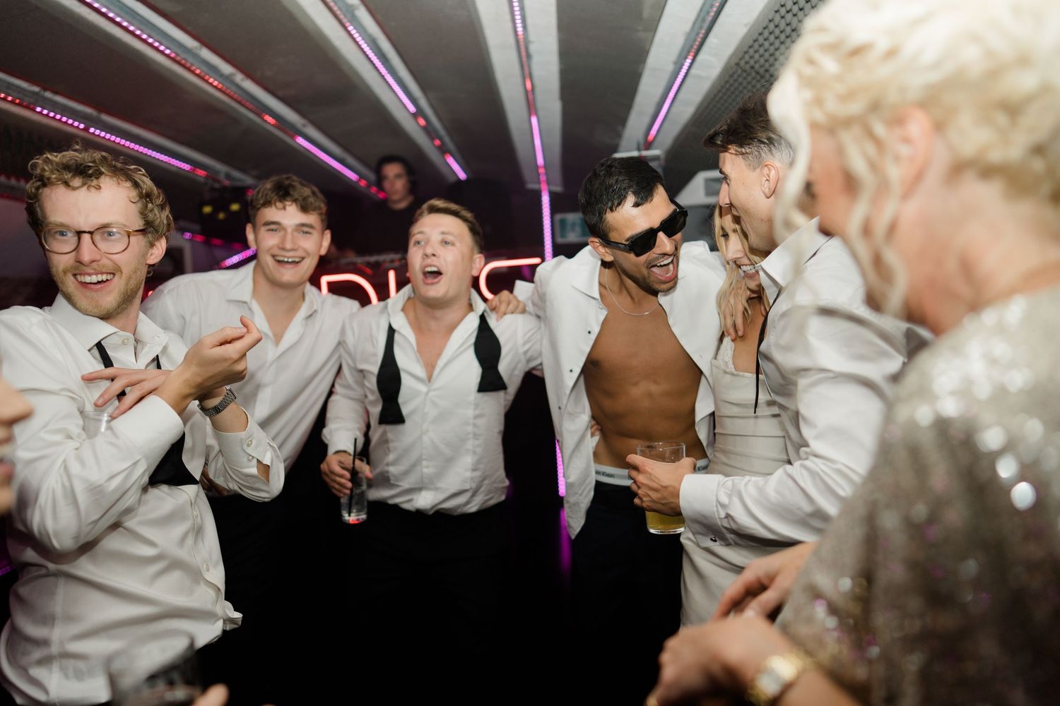 Groomsmen in white shirts celebrating and laughing together at a lively wedding party on a bus.