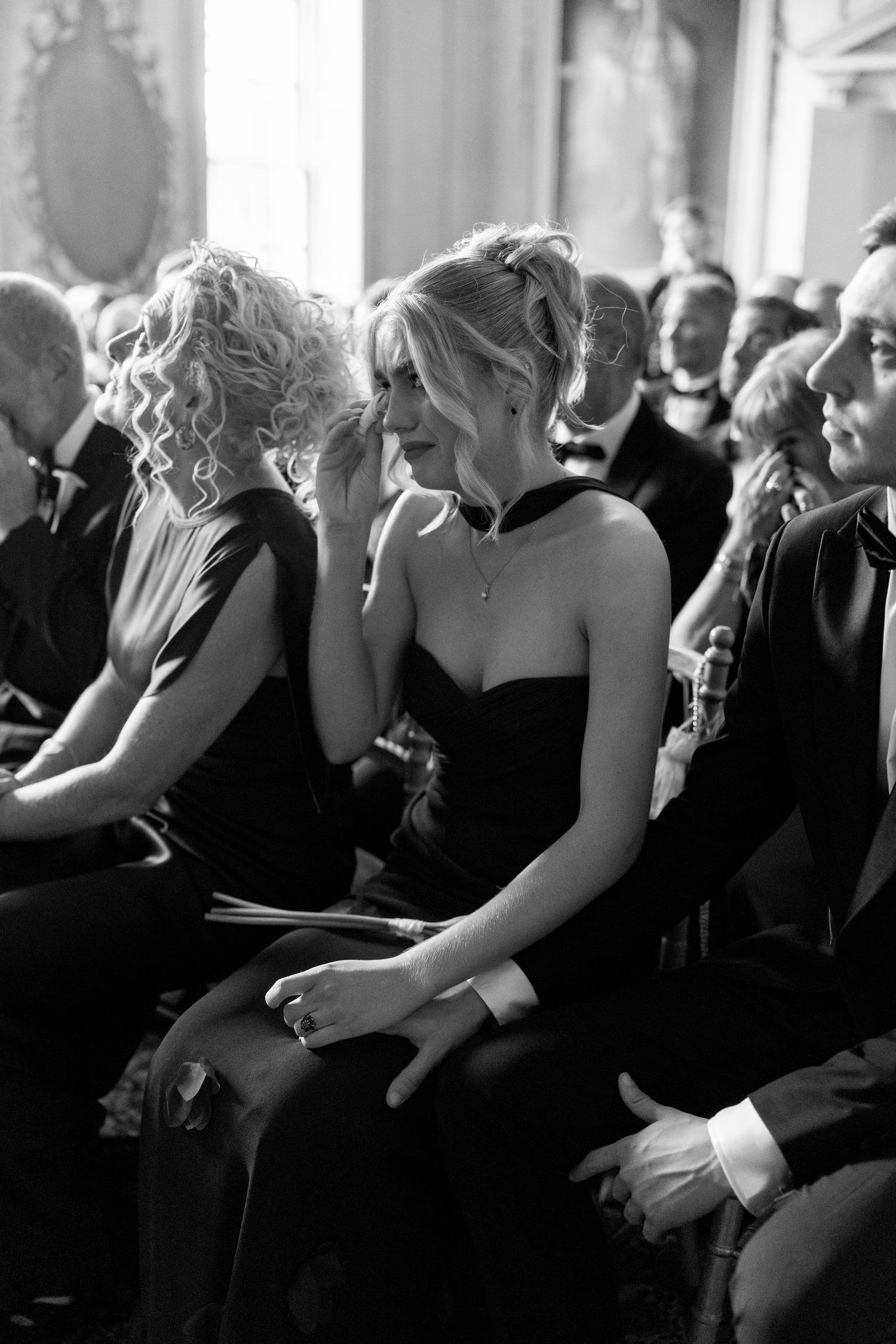 Emotional bridesmaid in black strapless dress wiping tears during wedding ceremony seated in pews.