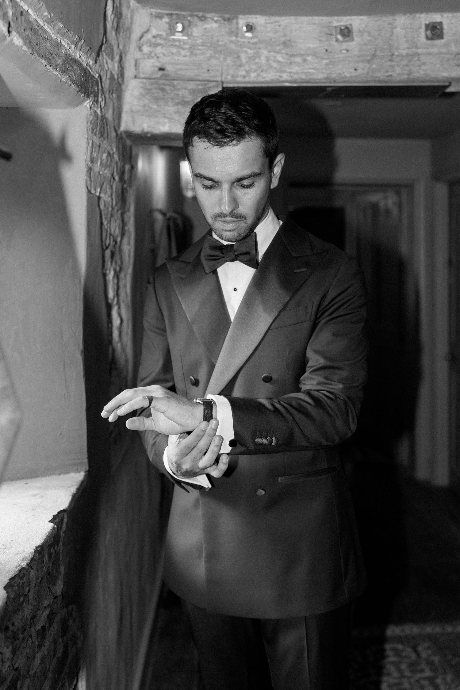 Dapper groom in black tuxedo and bow tie adjusting cufflinks in black and white portrait.