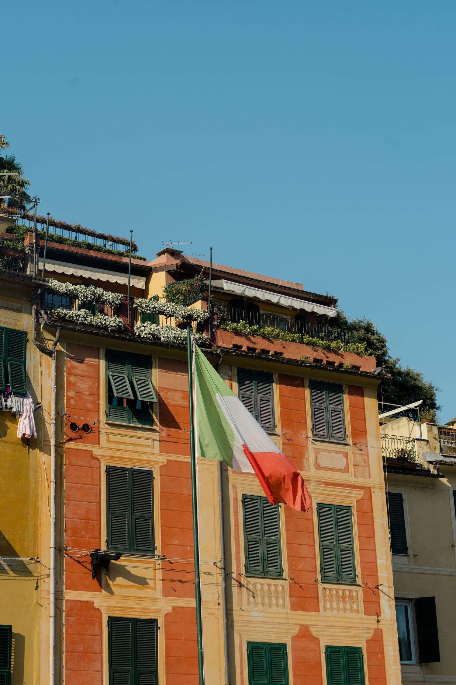 Italian flag waving on colorful orange building facade with green shutters and rooftop terrace under clear blue sky.