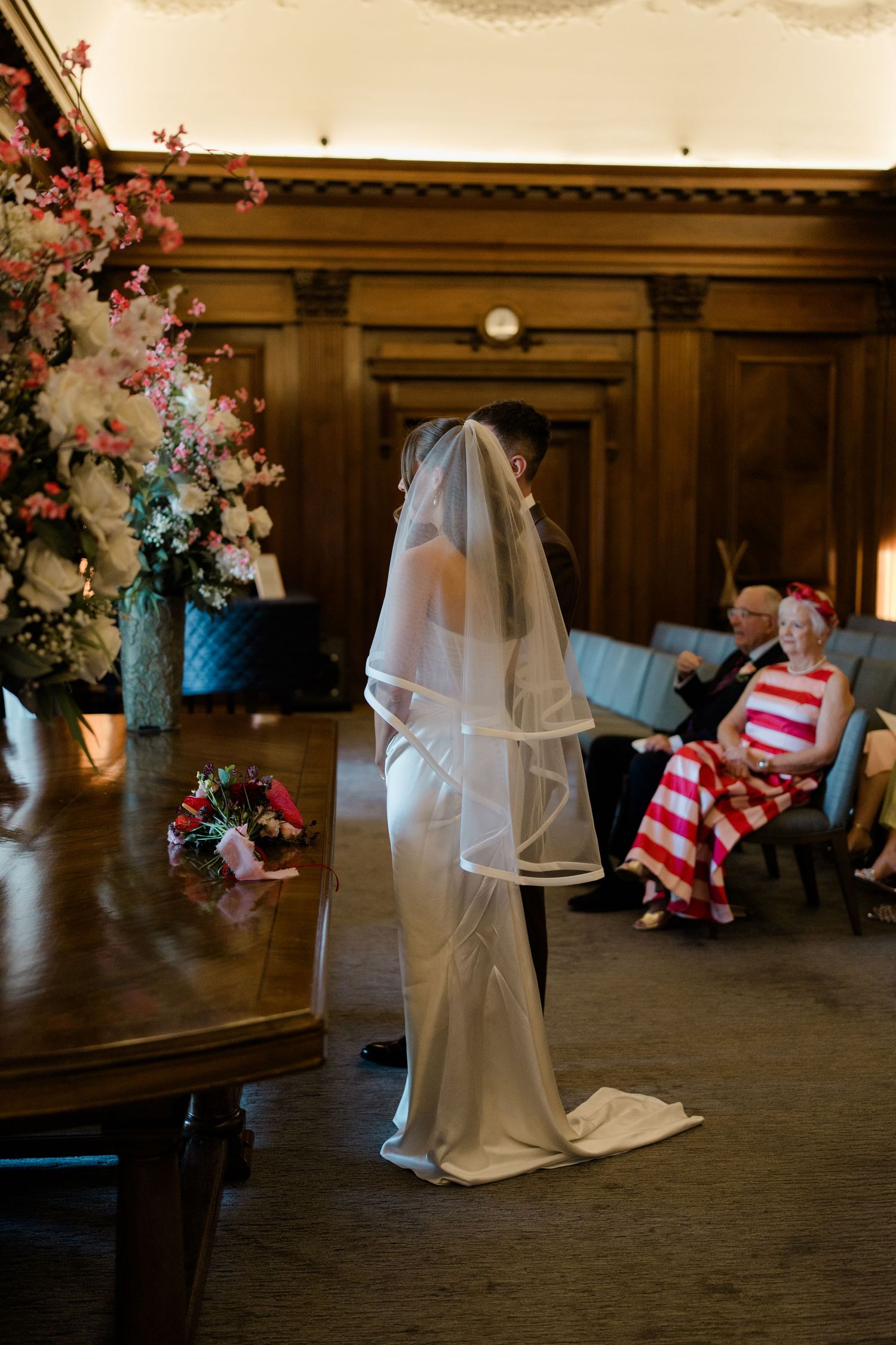 Bride in white gown and veil standing at the ceremony table in an elegant wood-paneled room.