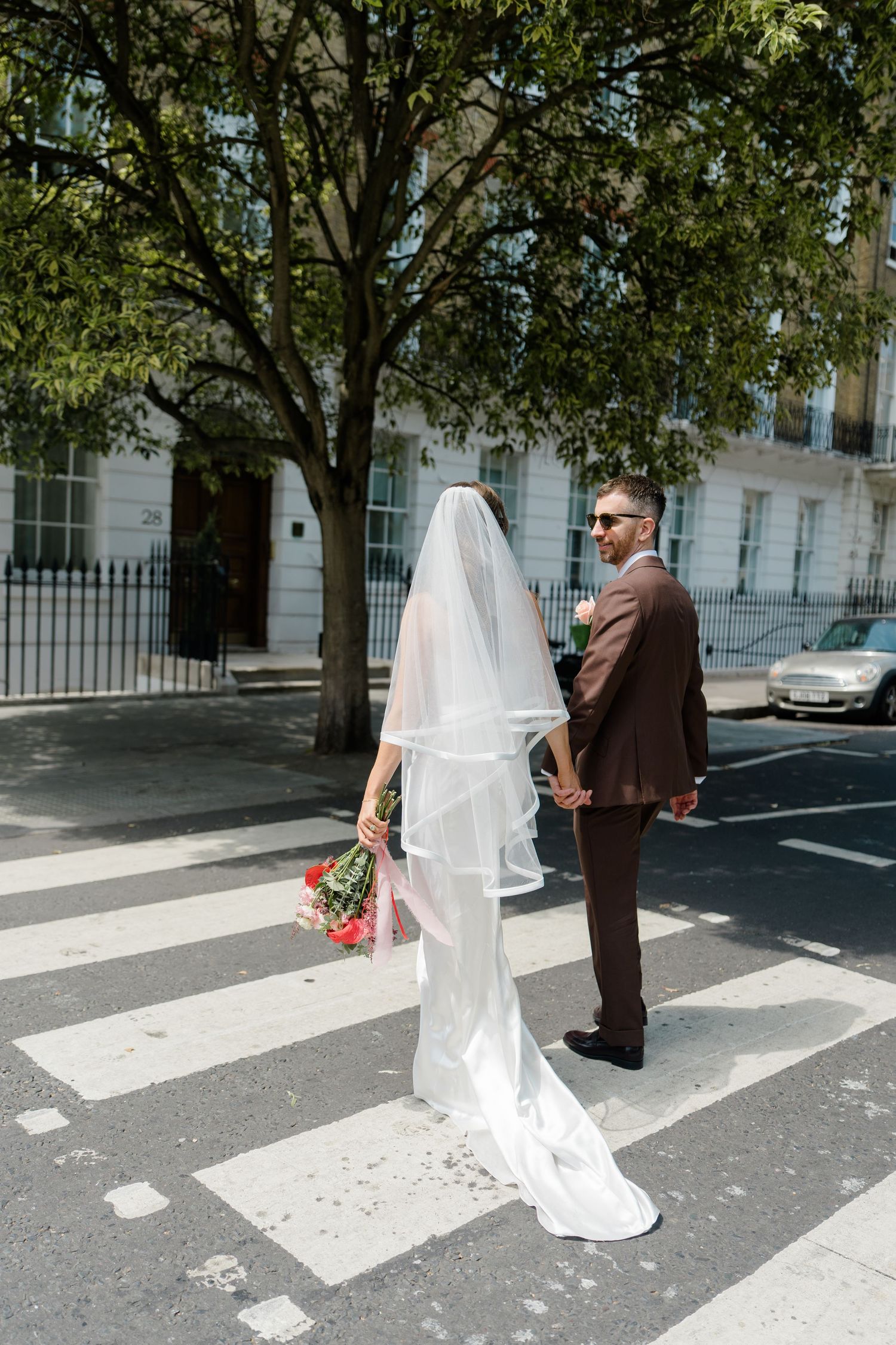 Bride in white gown and veil holds bouquet while crossing street with groom in brown suit on sunny day.