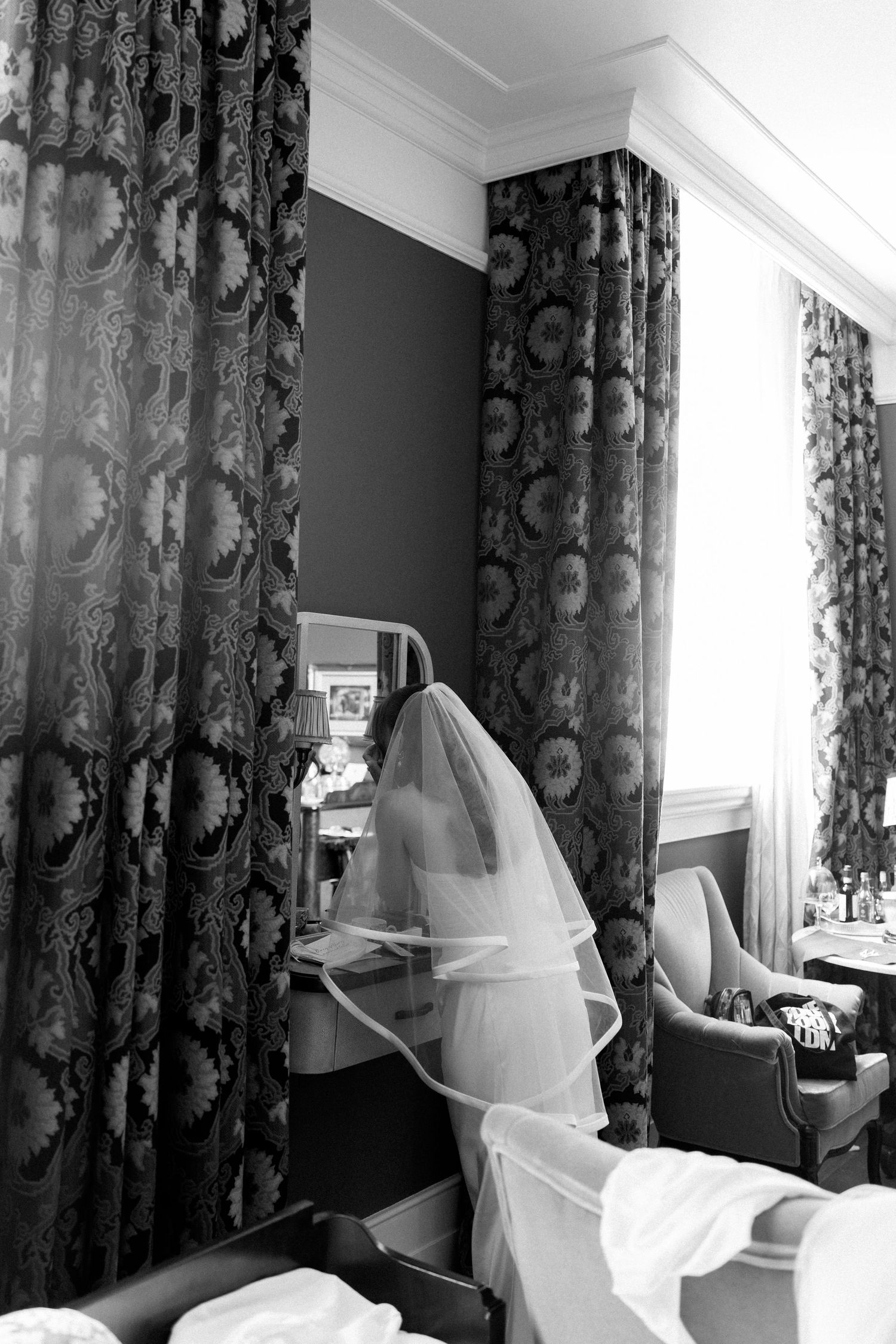 Bride in white wedding dress and veil standing by ornate curtains in elegant hotel room, black and white photo.