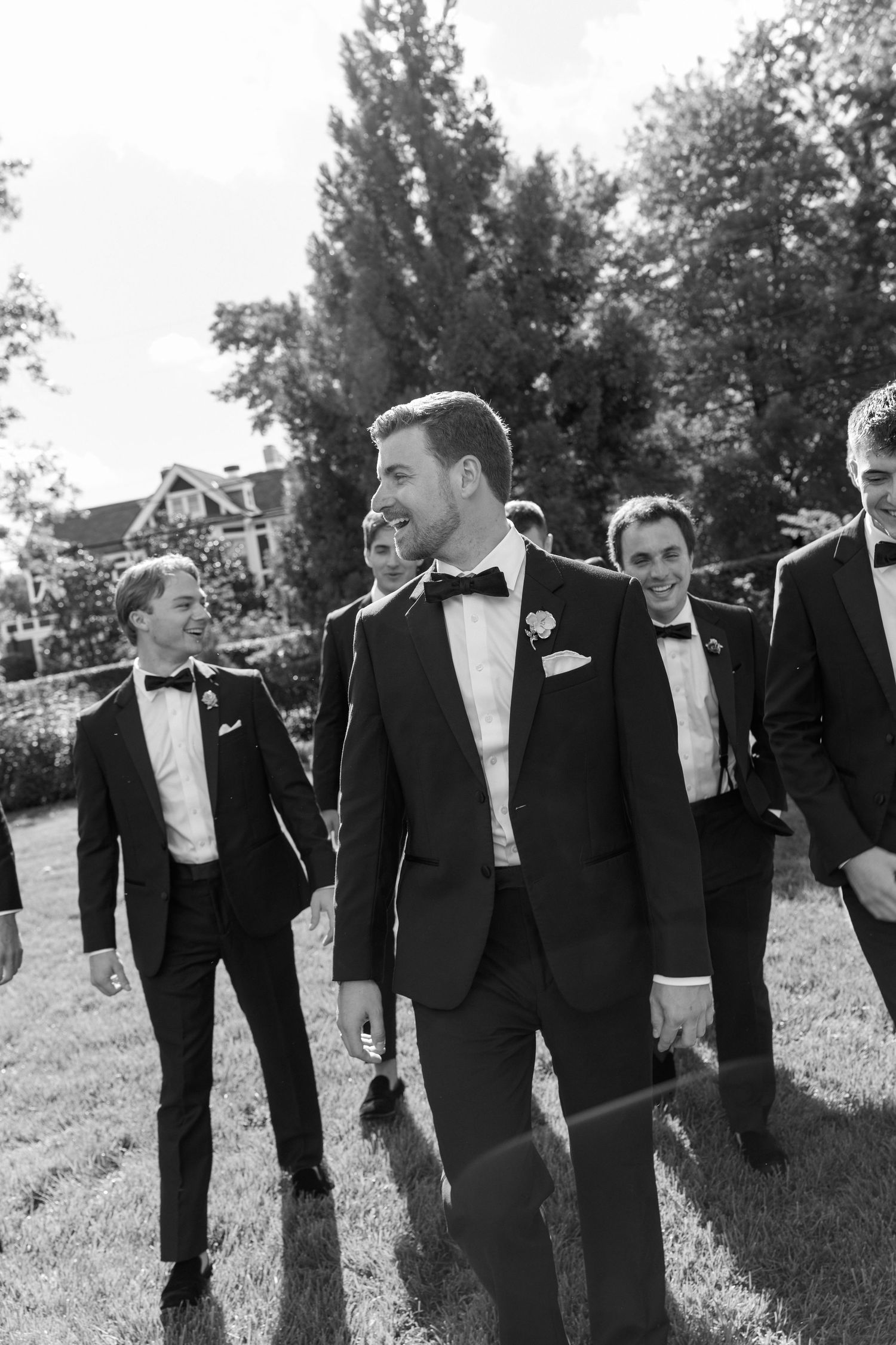 Groom and groomsmen in black tuxedos with bow ties walking outdoors on grass at a wedding celebration.