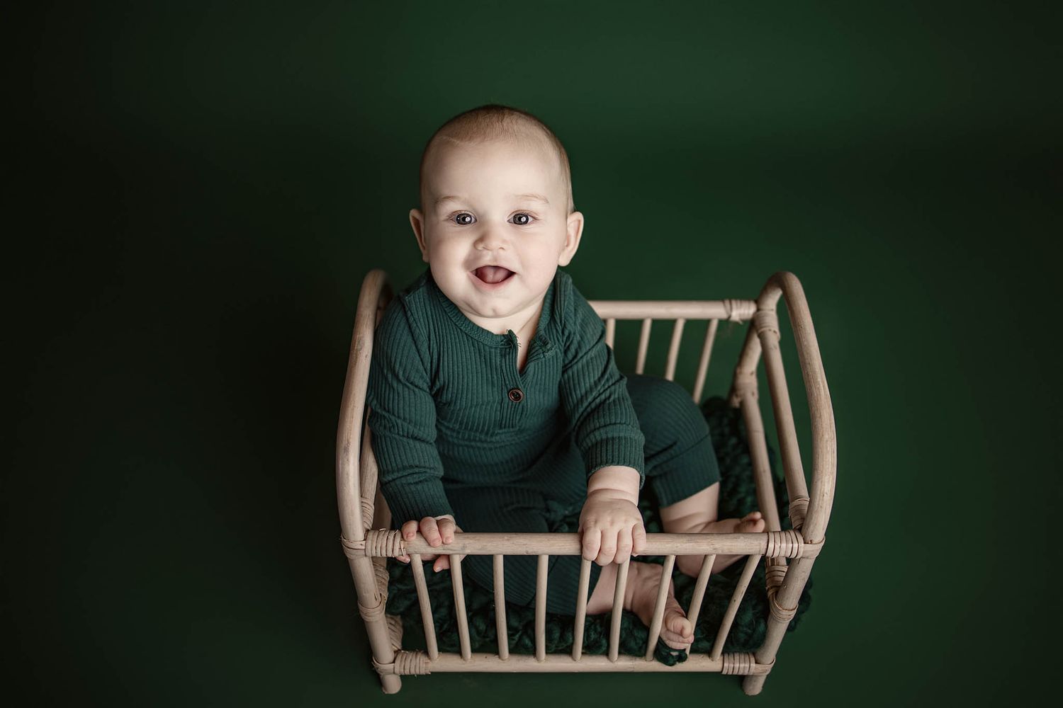 Baby exploring wooden crib against dark green backdrop.