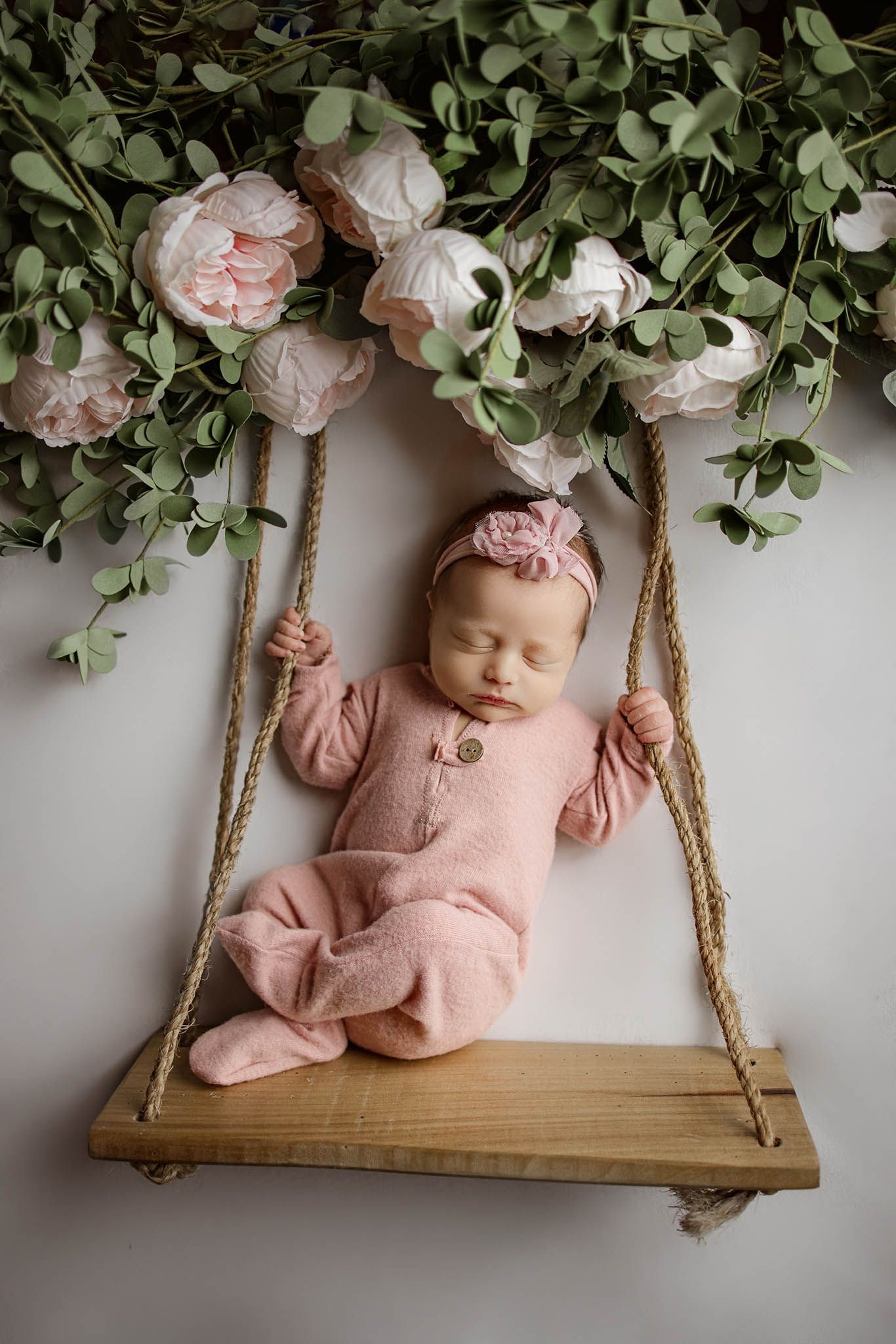 Newborn in pink outfit sleeping on wooden swing decorated with greenery and flowers.