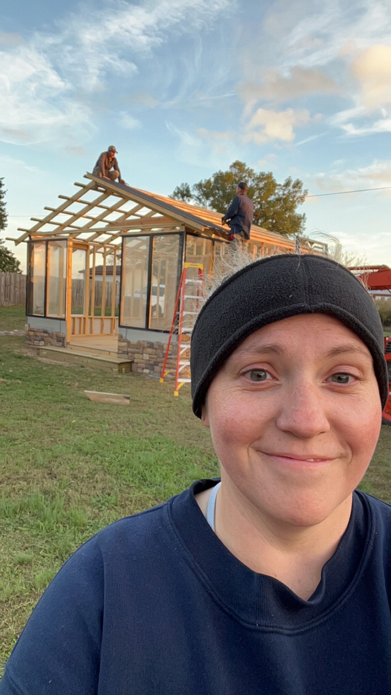 Construction site with wooden pergola structure and person in blue shirt and black hat taking series of selfie photos.