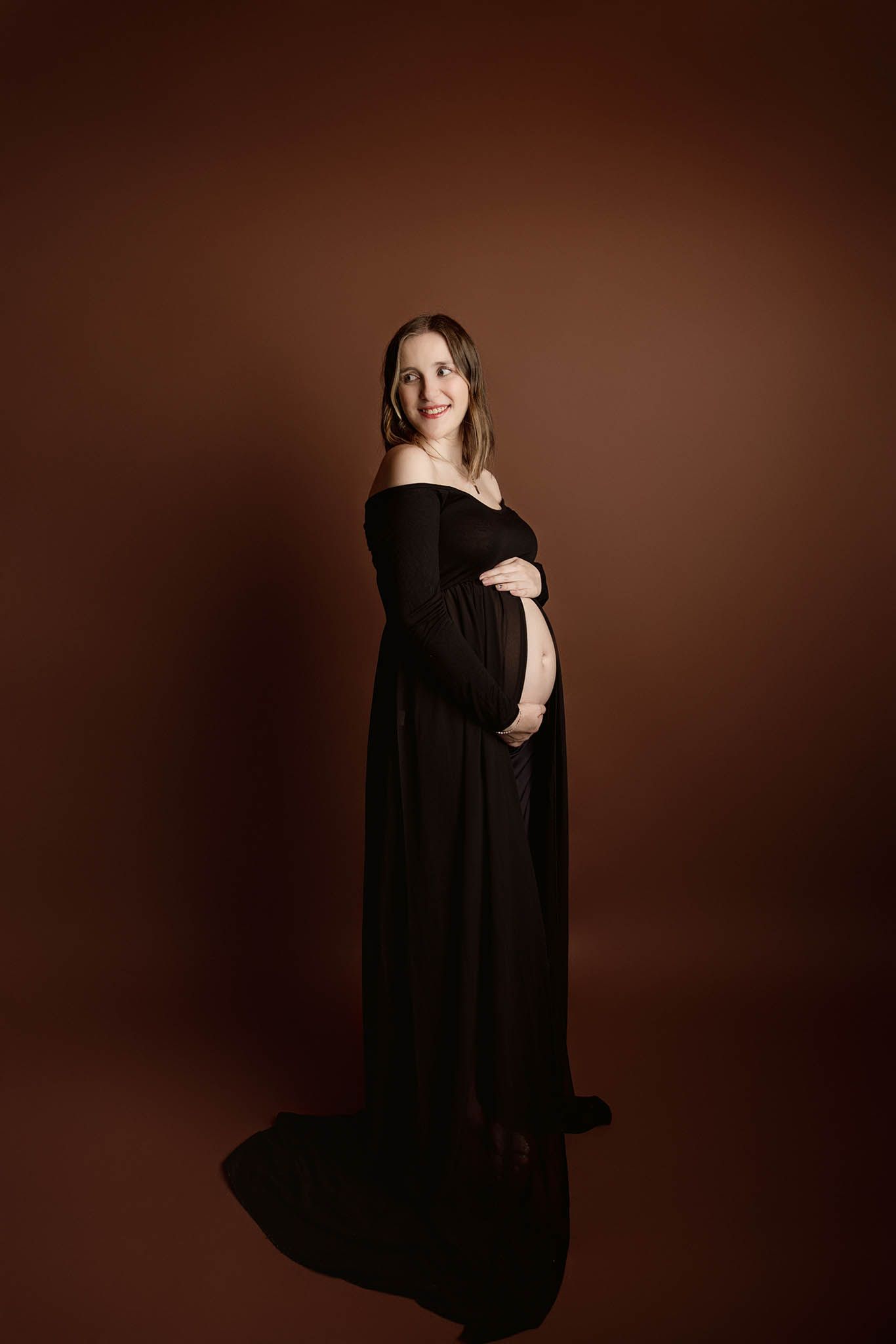 Elegant maternity portrait featuring an off-shoulder black gown against a dark brown studio backdrop.