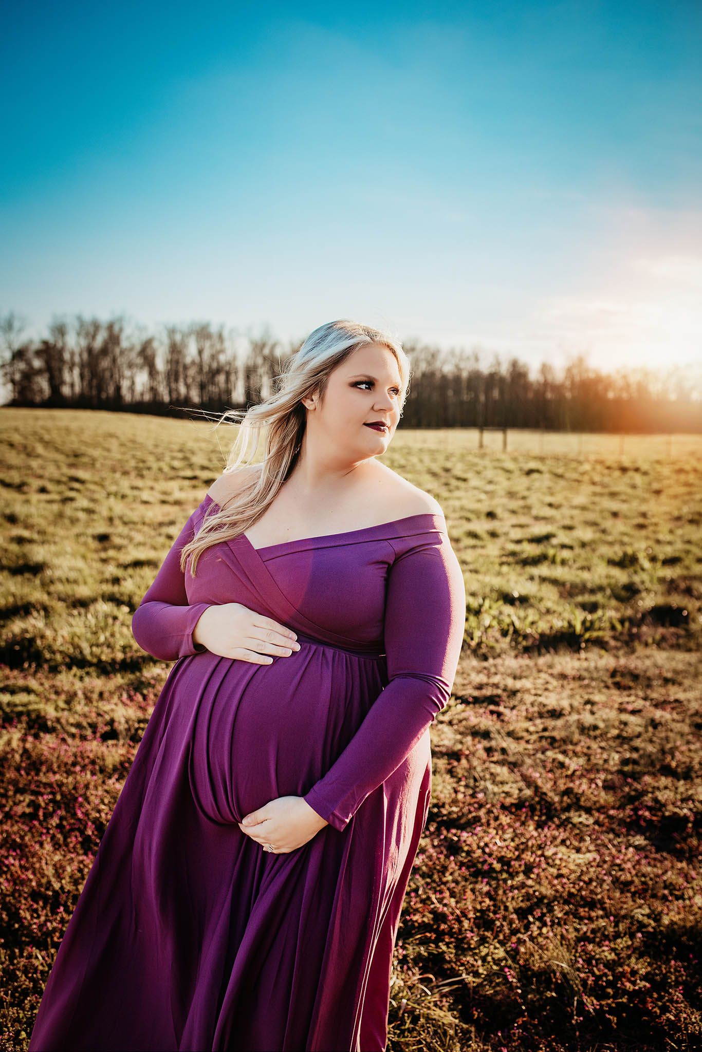 A purple maternity gown flows elegantly in a sun-lit field at golden hour, capturing a serene outdoor moment.