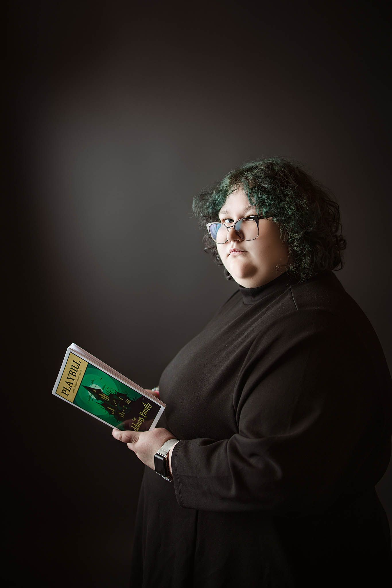 A figure in dark clothing reads a mysterious book against a black background in an artistic portrait series.