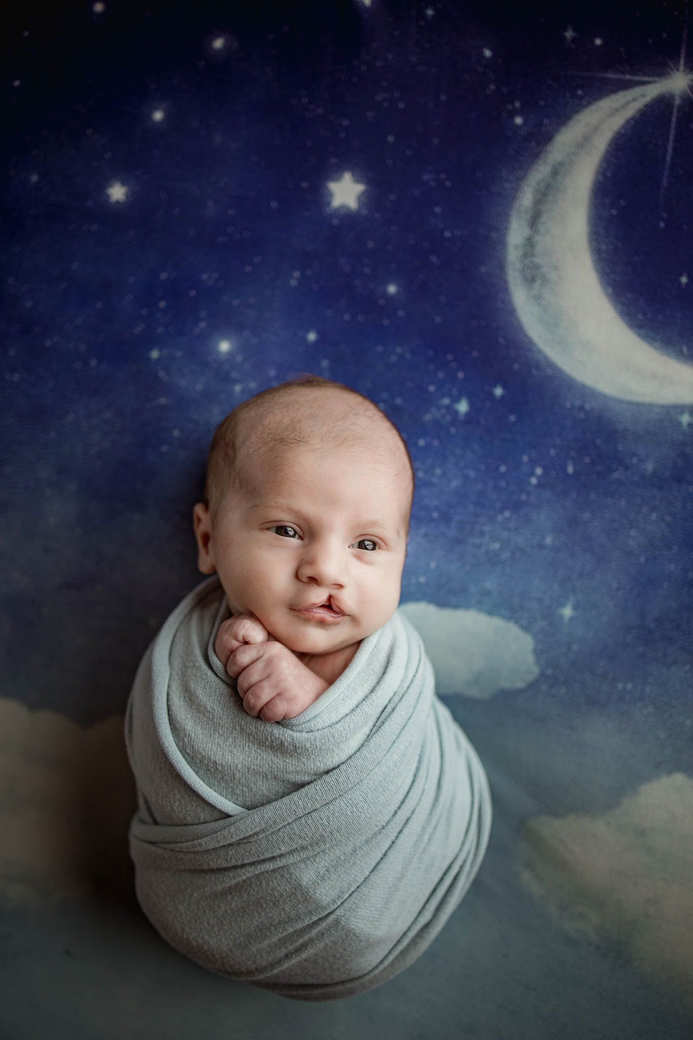Newborn wrapped in gray against starry night sky backdrop with crescent moon.