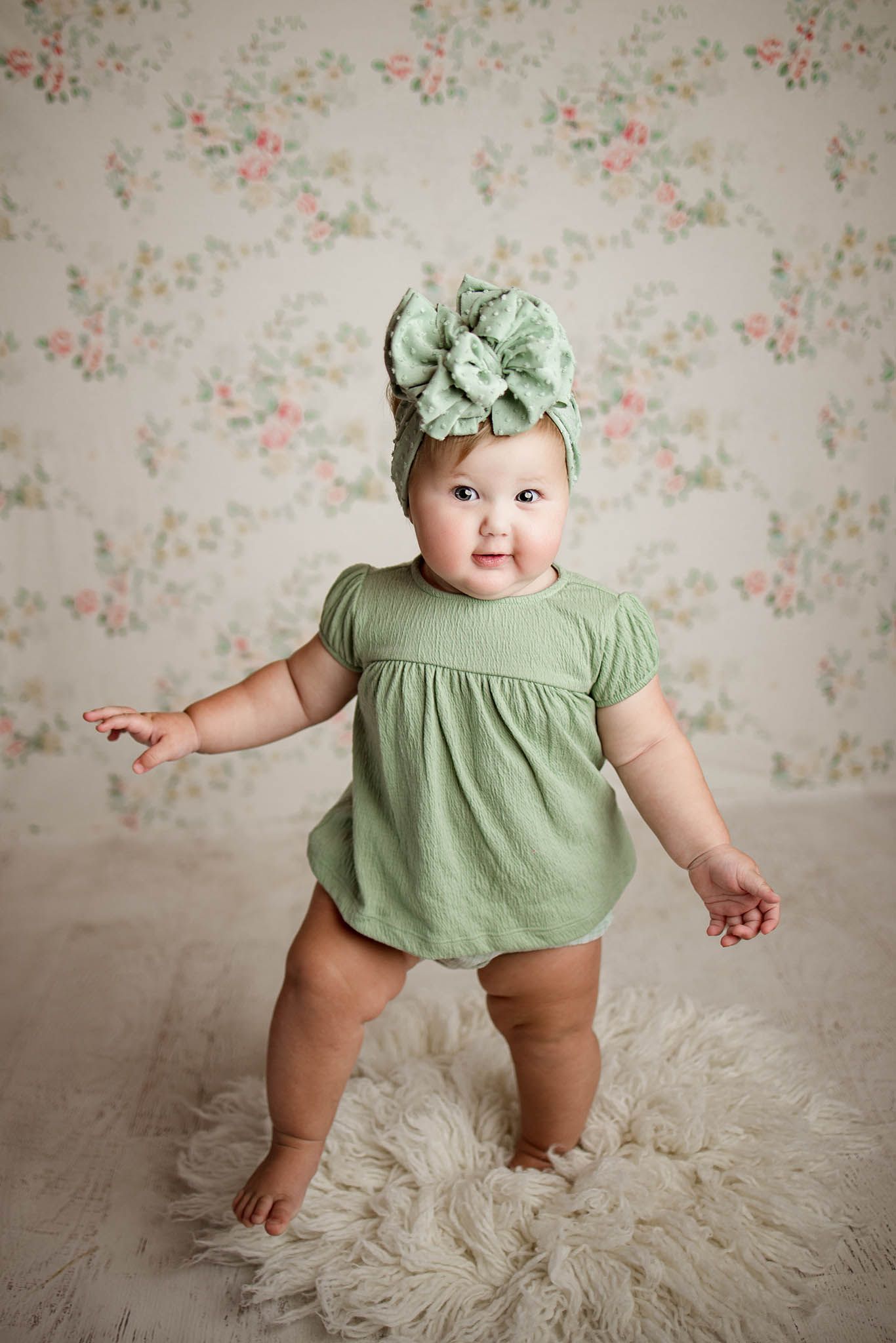 Baby wearing mint green dress poses against floral wallpaper on cream colored rug.