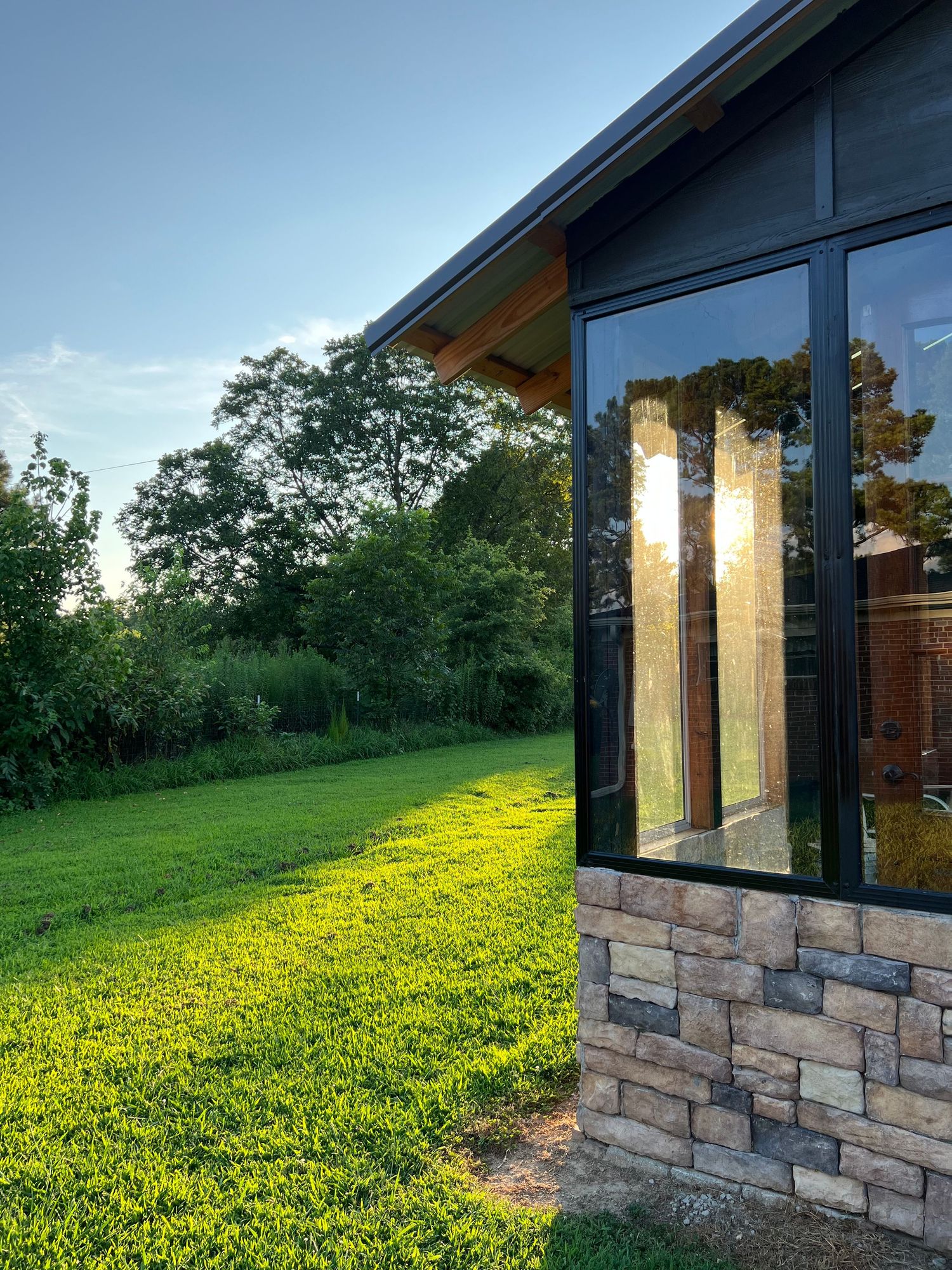 Modern house exterior with stone facade and tall windows overlooks a sunlit green lawn at golden hour.