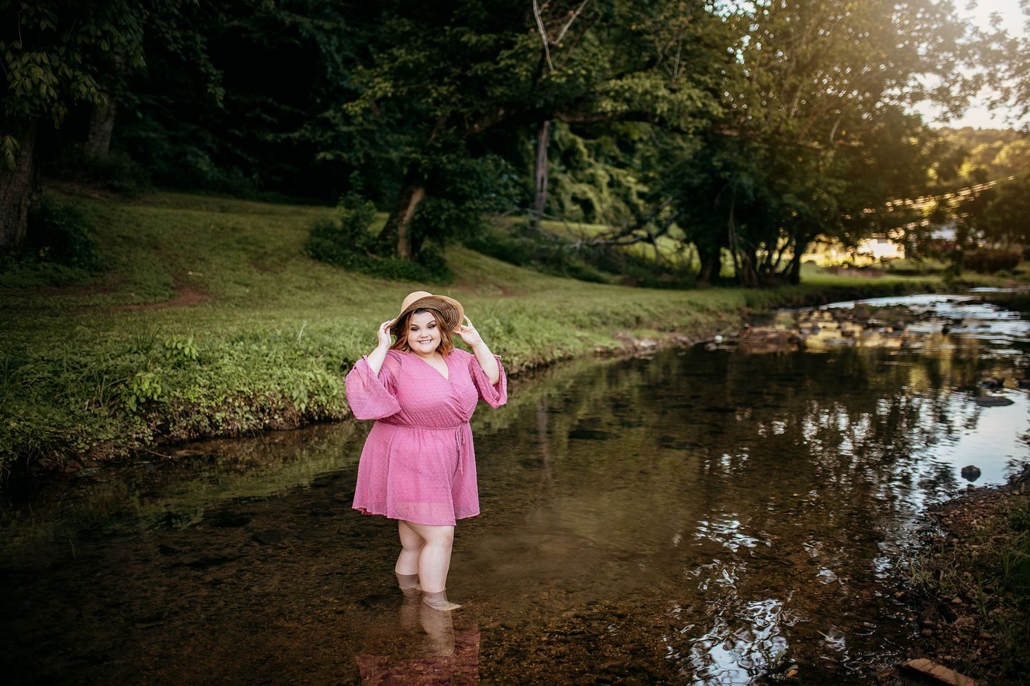 Young person in pink dress walking near stream in natural outdoor setting at sunset.