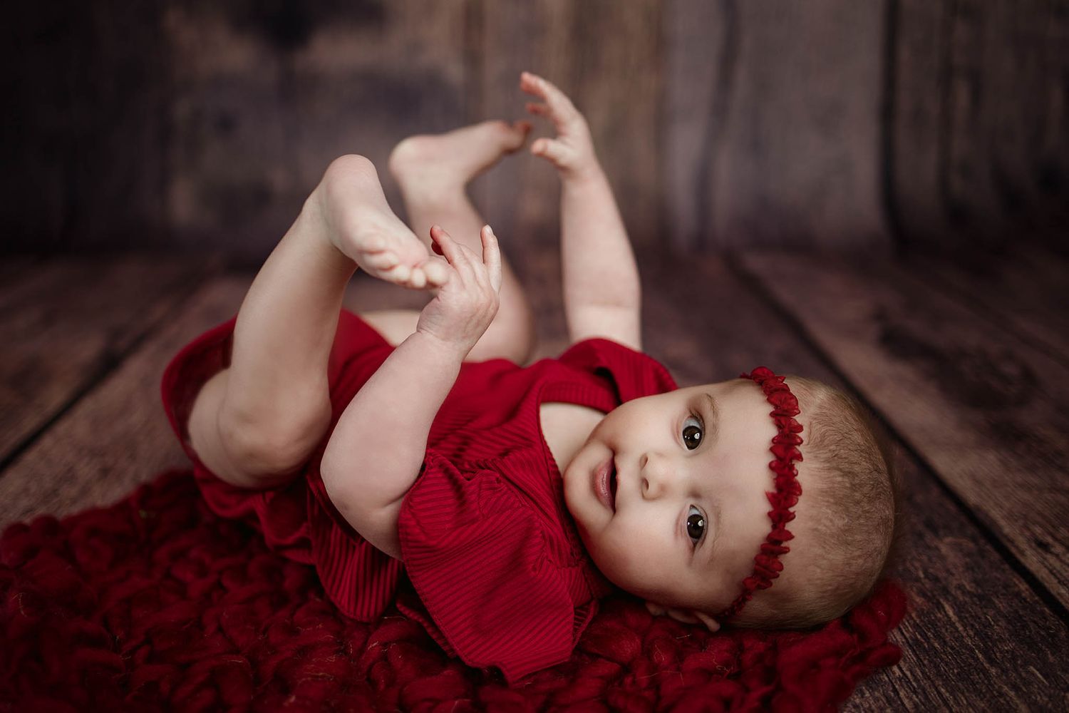 Baby in red festive outfit lying on dark wooden floor during photo session.
