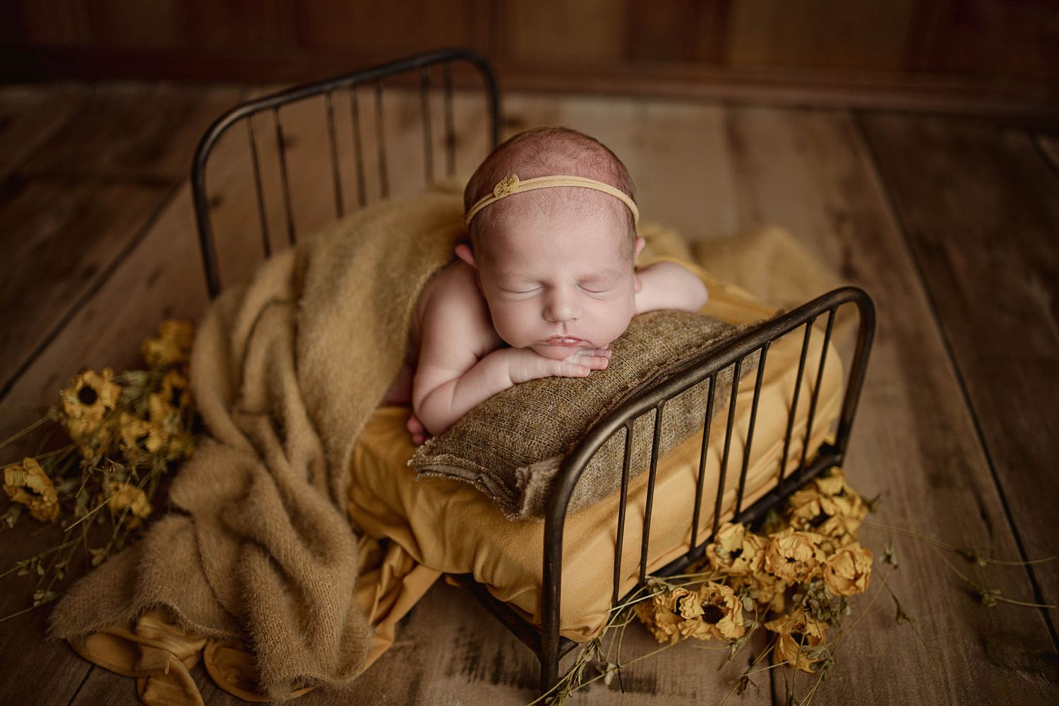 Newborn baby sleeping in vintage metal bed with yellow sunflowers and tan blanket in rustic setting.