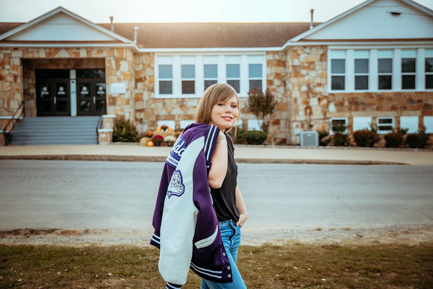 Student carries a surfboard while walking across parking lot in front of a stone building on an autumn day.