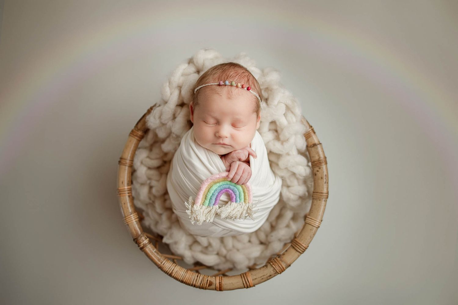 Sleeping newborn in white wrap surrounded by cloud-like cotton and rainbow accent.