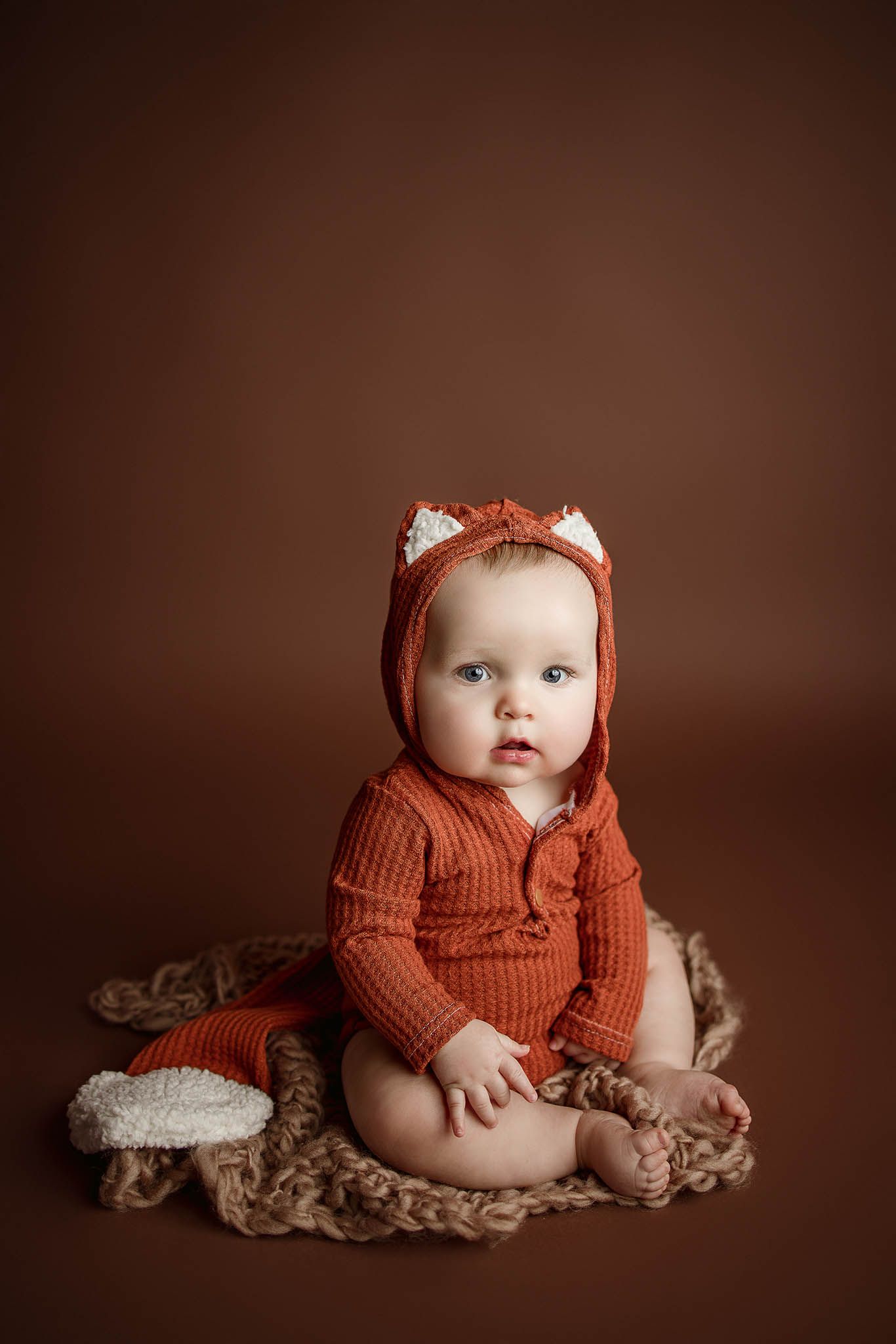 Baby wearing orange knit cardigan and cat ear headband on fuzzy brown blanket.