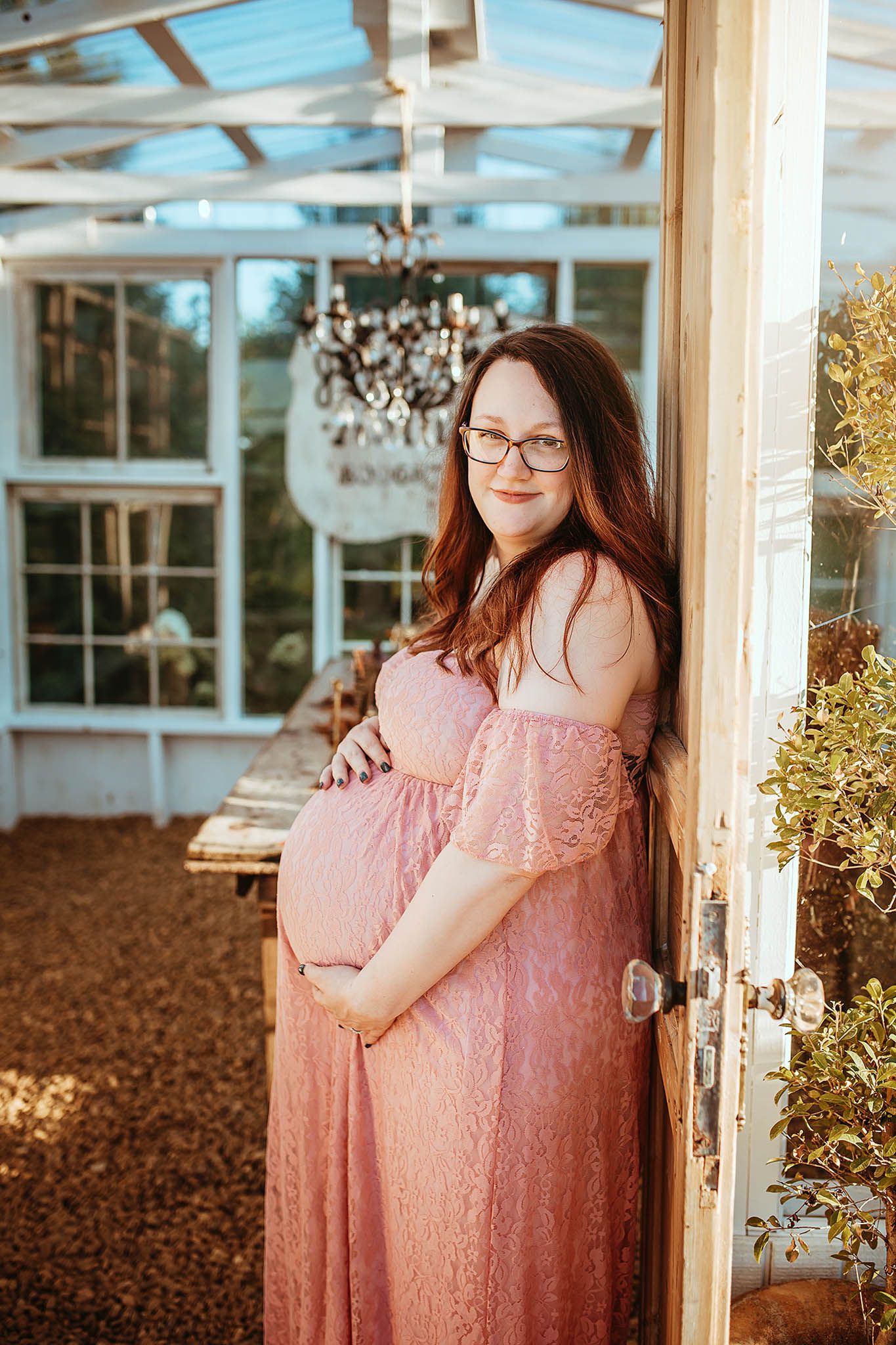 Maternity photoshoot in a rustic greenhouse setting with soft pink lace dress and natural lighting.
