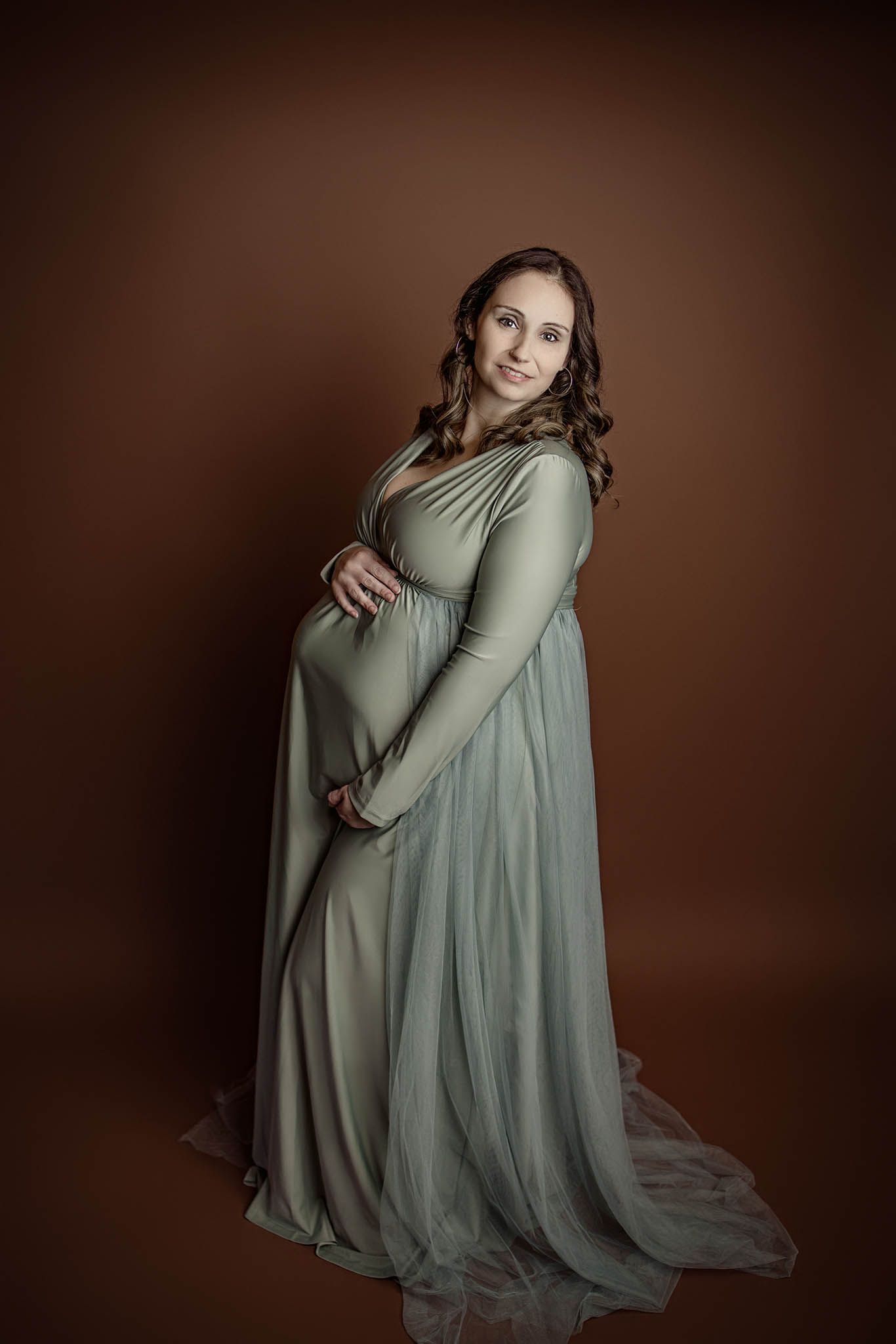 Mother in a flowing sage green dress poses gracefully against a dark brown backdrop in an artistic portrait series.