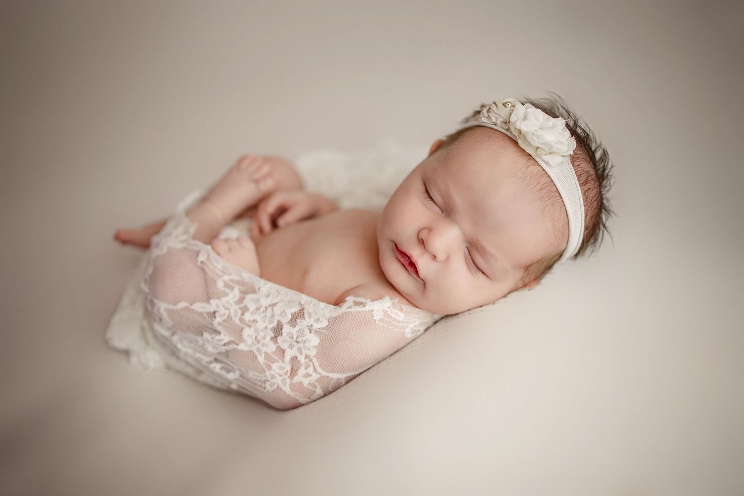 Newborn baby sleeping peacefully wrapped in delicate white lace on neutral background.