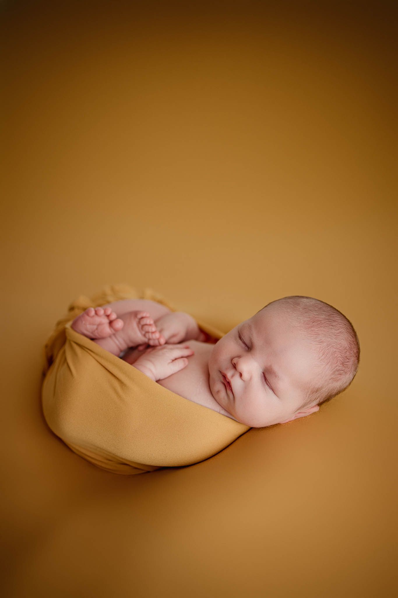 Sleeping newborn wrapped in golden fabric against warm brown background.