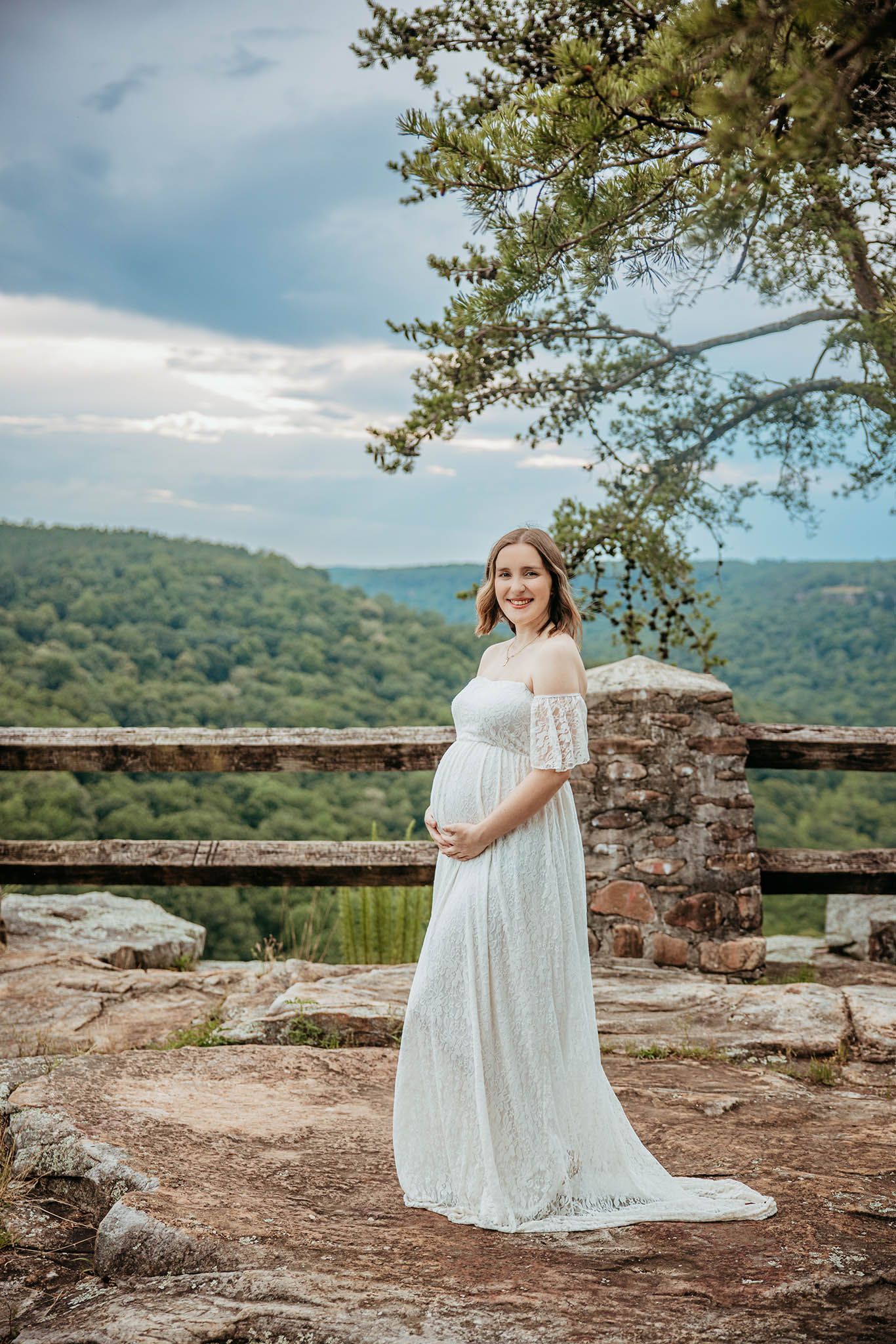 Maternity photo series of woman in flowing white lace dress posed against stone wall with mountain landscape backdrop.
