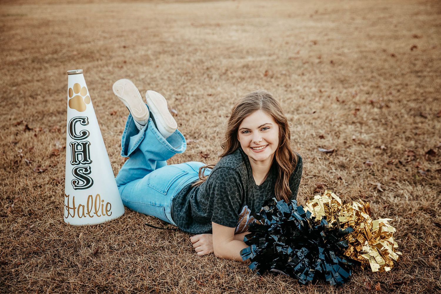 A cheerleader in blue uniform sits in fall field holding poms and spirit megaphone while smiling towards camera.
