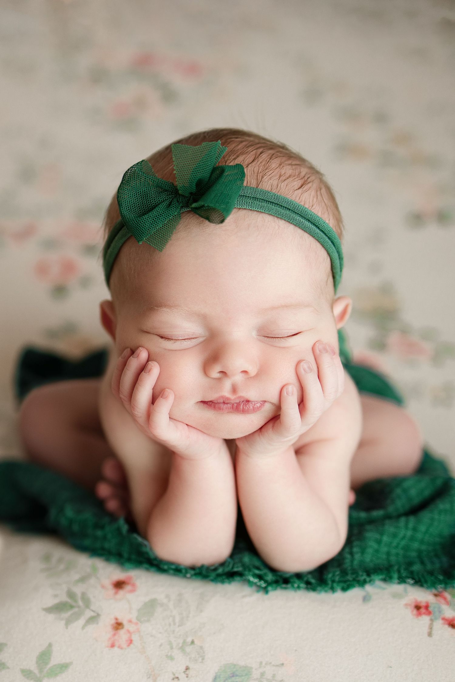 Smiling newborn in green headband resting head on hands against floral backdrop.