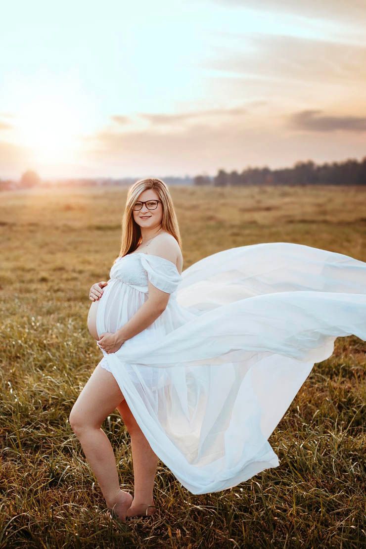 Ethereal maternity photo in flowing white dress at sunset in golden field.