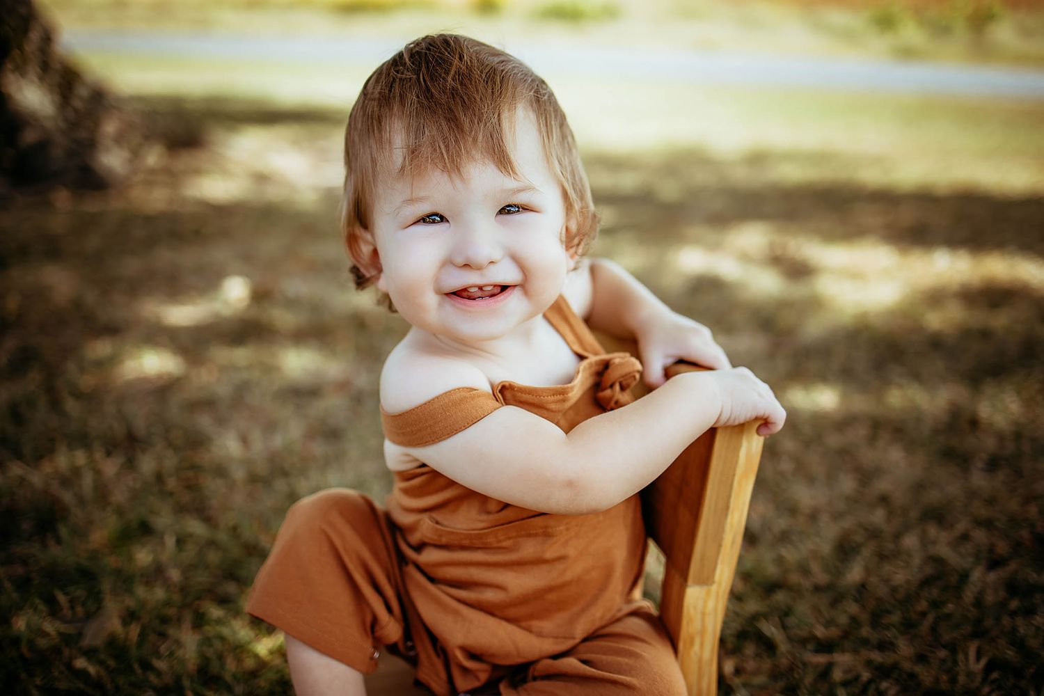 Happy baby sits outdoors in brown outfit on wooden chair with natural lighting.