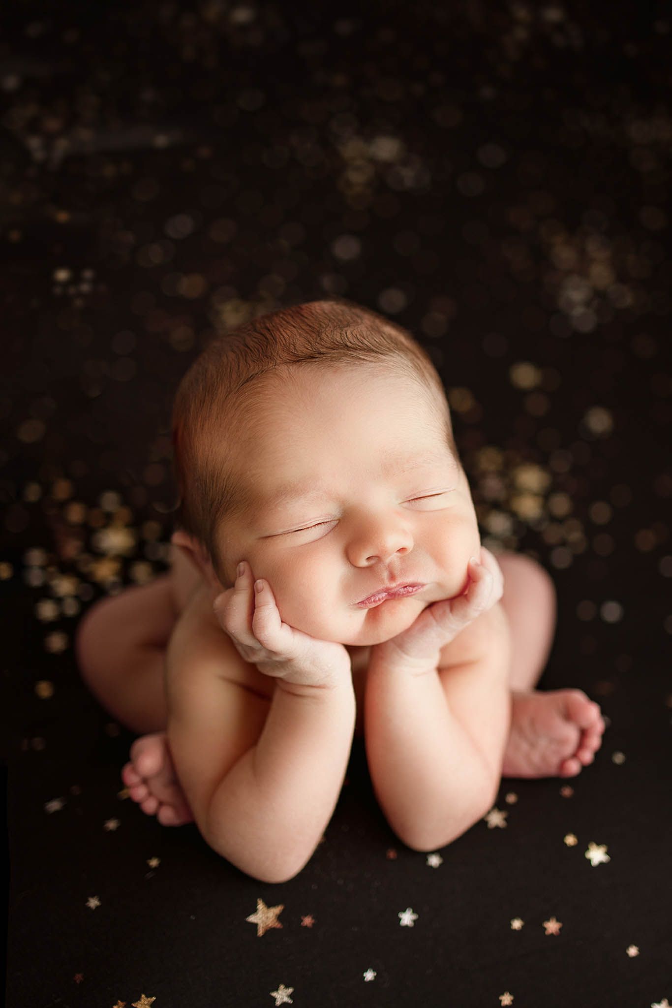 Sleeping newborn posed on dark backdrop with scattered golden star decorations.