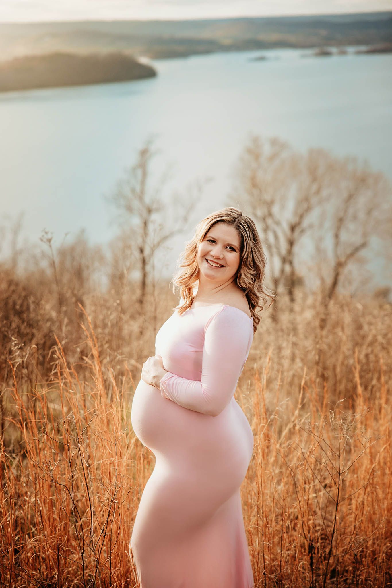 Expectant mother in soft pink dress stands among golden autumn grasses near a serene lake at sunset.