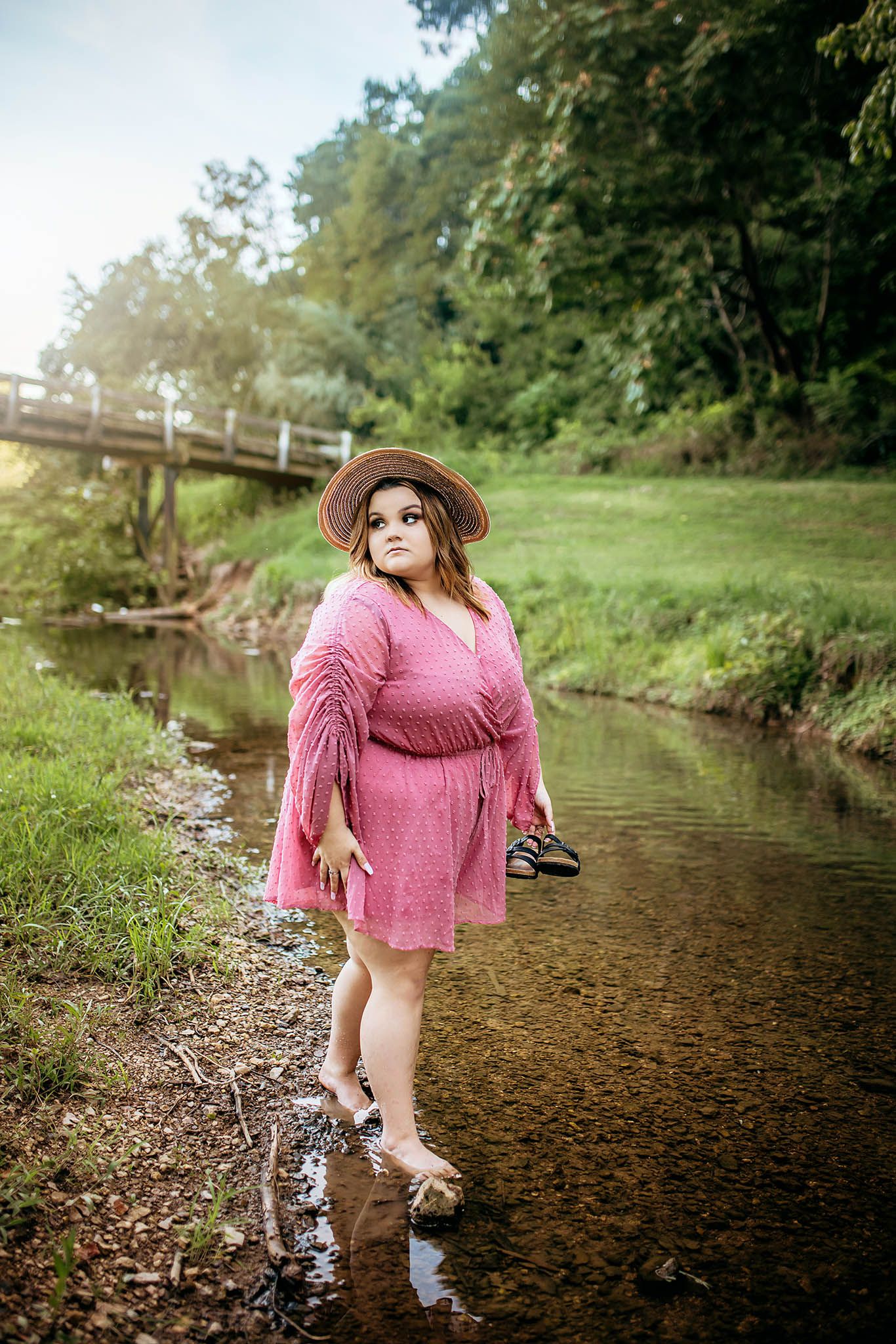 Person in pink dress playing in shallow creek water surrounded by green foliage.