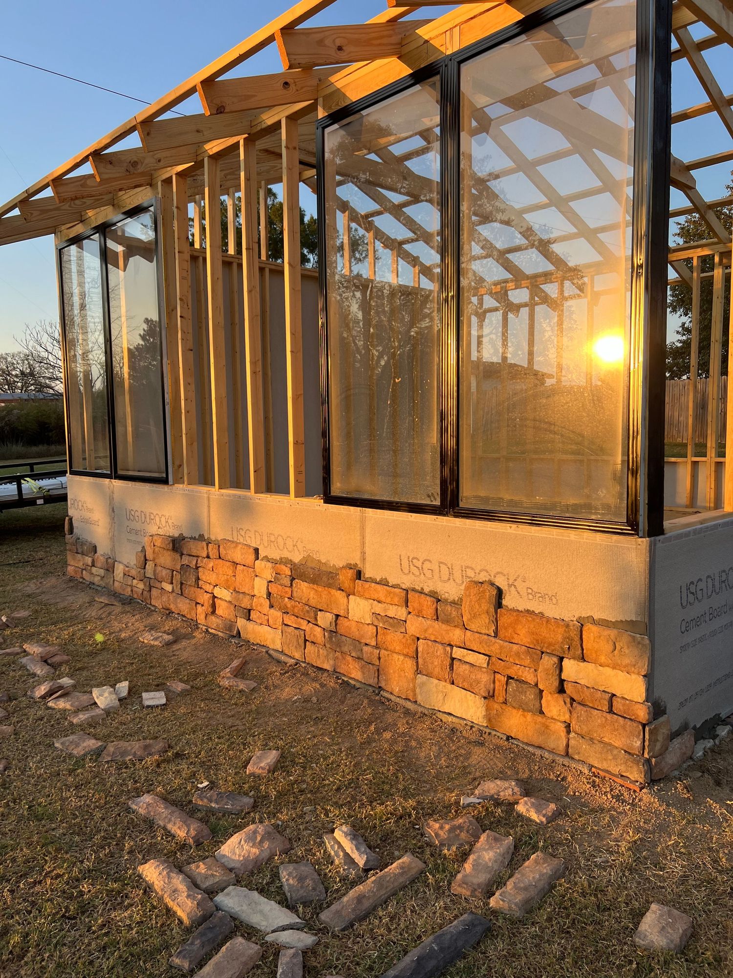 Greenhouse or sunroom under construction with stone base foundation illuminated by golden sunset light through glass panels.