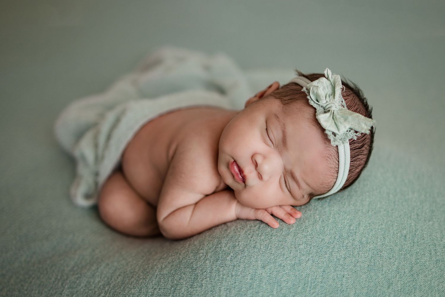 Peaceful sleeping newborn wrapped in soft sage green fabric with delicate white headband.