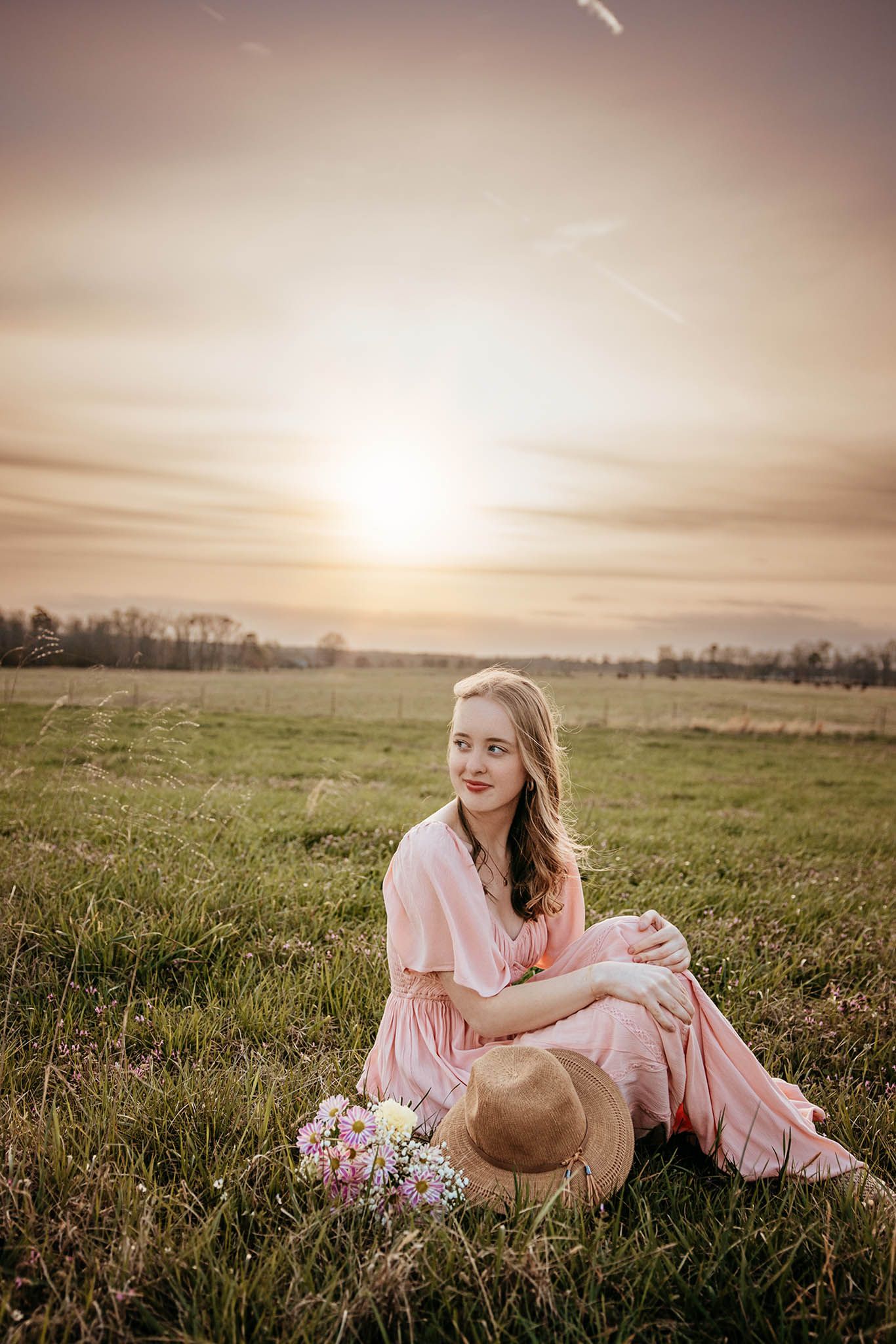 A romantic sunset portrait in a grassy field with a pink dress and straw hat against a golden sky.