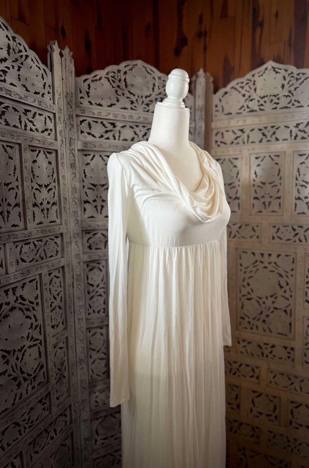 Elegant white flowing wedding dress with draped neckline displayed against ornate carved wooden screen backdrop.
