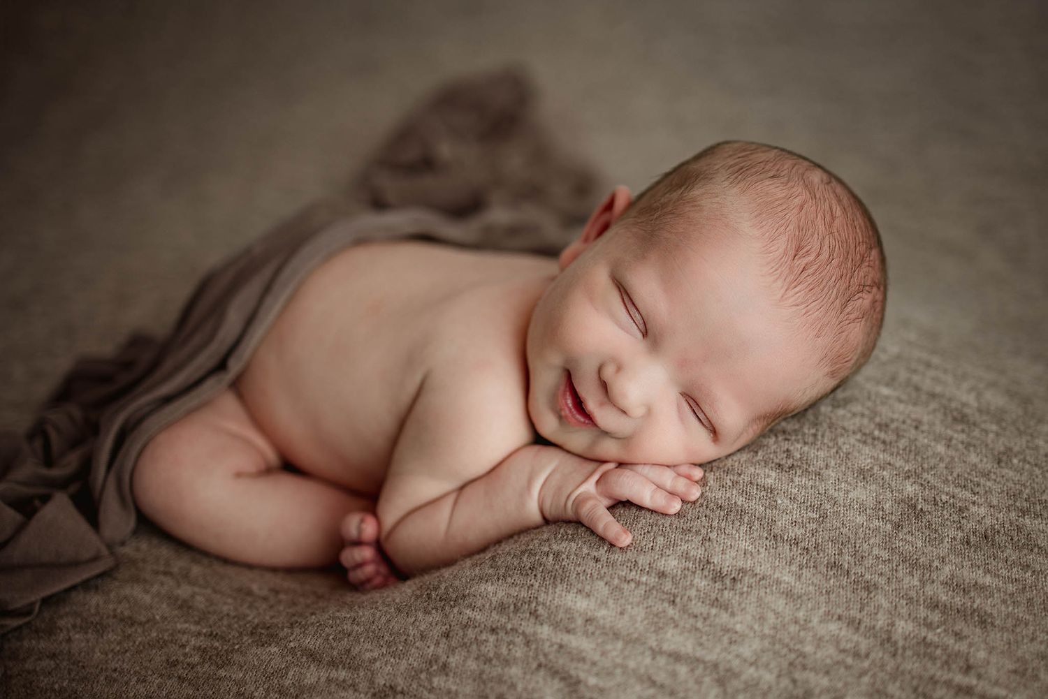 Sleeping newborn curled up on textured gray fabric in natural light.